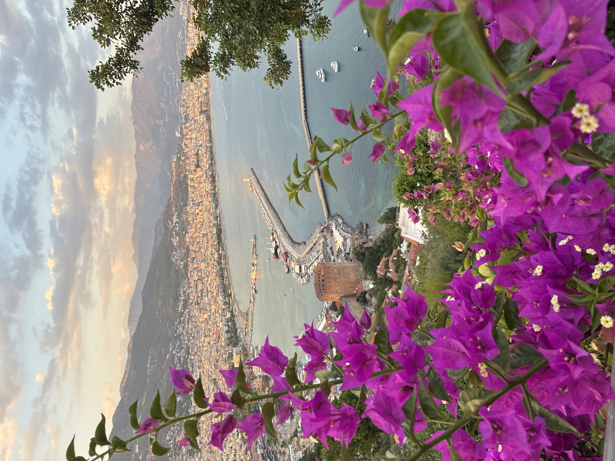 The image captures a scenic view of Alanya, Turkey, from an elevated vantage point, likely a hill overlooking the city and the Mediterranean Sea. In the foreground, vibrant purple bougainvillea flowers are in full bloom, their petals and green leaves creating a soft, colorful frame for the scene beyond.
The city of Alanya stretches along the coastline, its buildings a mix of light-colored structures with red-tiled roofs, bathed in the warm, golden light of what appears to be late afternoon or early evening. The coastline curves gently, forming a bay filled with boats and a prominent pier.
Dominating a section of the coastline is a historical red-brick tower, likely a part of Alanya's castle or fortifications. A long, man-made pier extends into the sea, featuring a lighthouse at its end. Several boats, including yachts and smaller vessels, are anchored in the bay or navigating the calm waters.
In the background, a range of imposing mountains forms a dramatic backdrop to the coastal city, their slopes covered in lush green vegetation. The sky is a canvas of soft, pastel colors, with scattered clouds tinged by the setting sun. The overall atmosphere is tranquil and picturesque, showcasing the natural beauty and historical charm of Alanya.