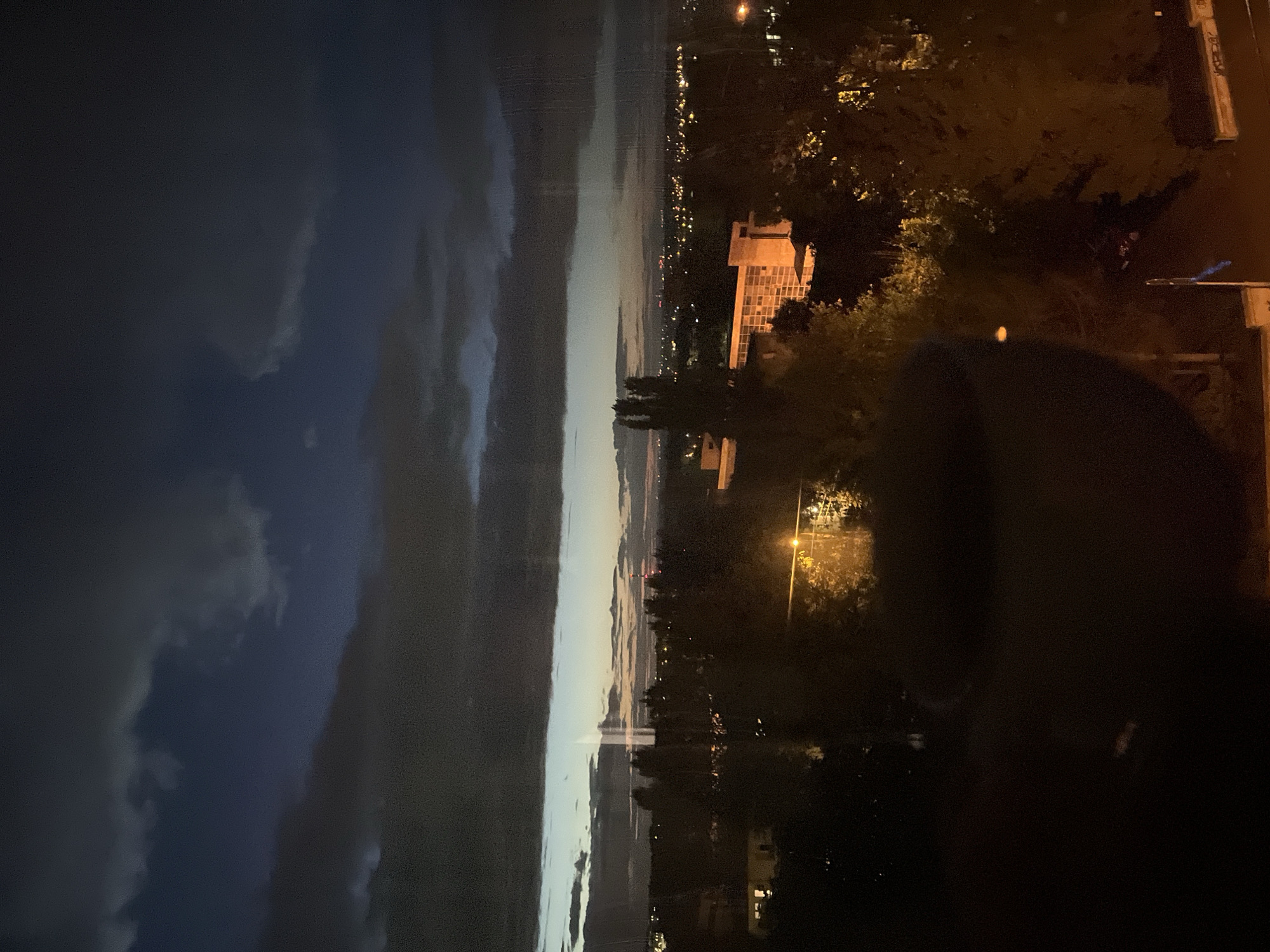 The image depicts a twilight cityscape viewed from an elevated position, possibly a window, given the slight reflection on the left side. The sky is a gradient of dark blues and grays, with scattered clouds casting shadows. A strip of lighter sky is visible near the horizon, suggesting the last vestiges of daylight. Below the sky, a dense arrangement of trees and buildings creates a dark, textured landscape. In the middle ground, a distinct building with illuminated windows and a light-colored facade stands out. Further in the distance, city lights twinkle, indicating a populated urban area. A streetlamp with a bright yellow light is visible in the foreground, illuminating the surrounding trees. The overall atmosphere is calm and serene, characteristic of the transition from day to night.