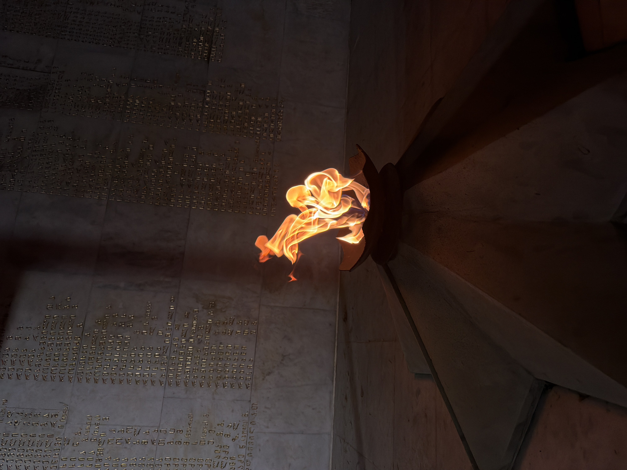 A close-up shot captures a burning eternal flame within a memorial wall, likely situated in Tbilisi, Georgia. The flame emerges from a dark metal fixture on a wall with chiseled inscriptions in Georgian, possibly memorializing individuals. The right side of the scene includes a corner wall, creating deep shadows. The overall lighting is dim, focusing attention on the flame's intense glow.