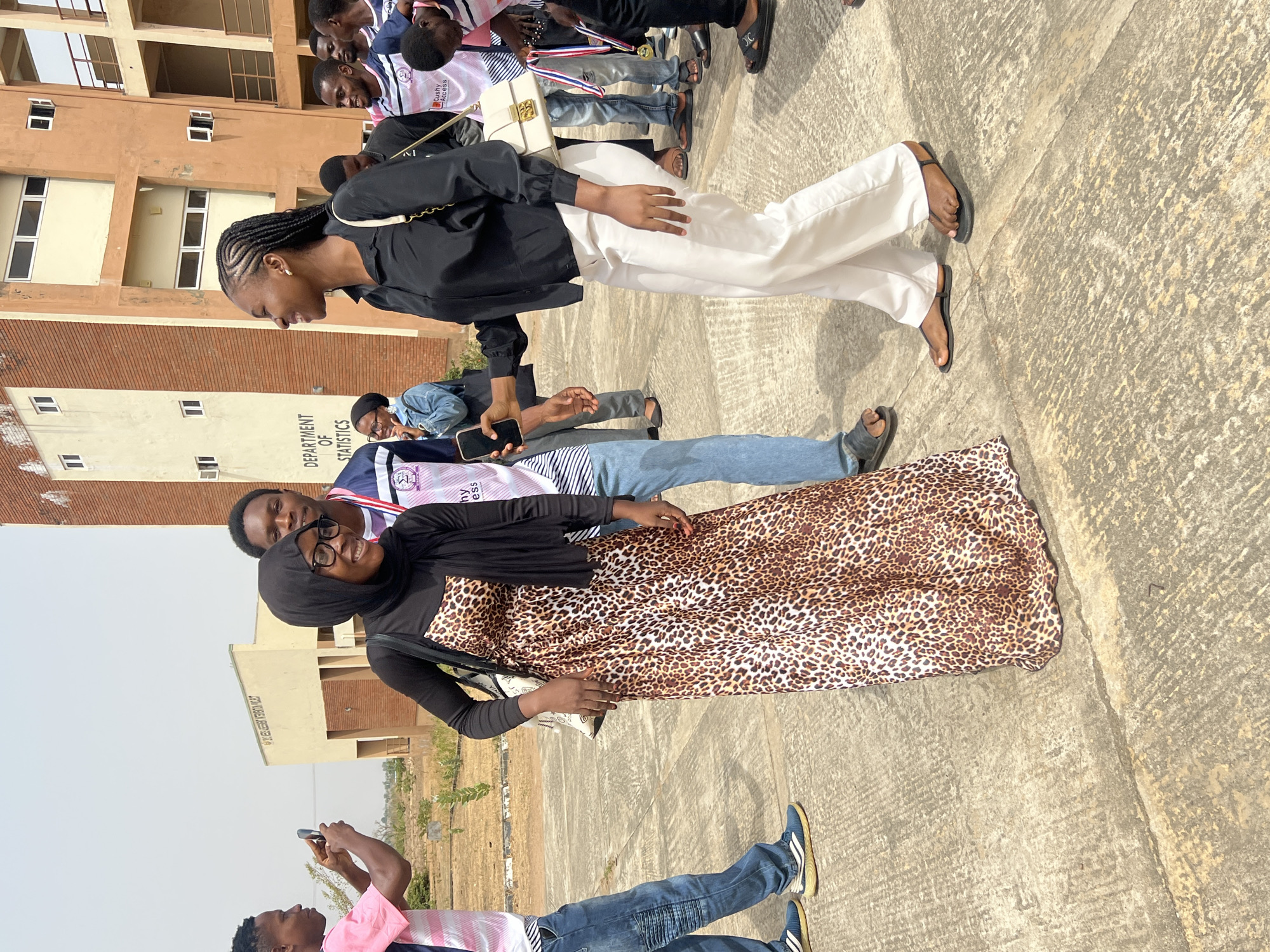 The image depicts several people in an outdoor setting. A woman wearing a black hijab, glasses, and a leopard print dress is in the foreground. Behind her is another woman wearing a black shirt and white pants. Several other people are visible in the background, some wearing shirts with logos. One person is holding up a phone, possibly taking a picture. The background includes a building with the words "DEPARTMENT OF STATISTICS" visible on its facade. The location is likely in Minna, Nigeria, given the building style and context of the photo. The environment appears to be a campus or public space. The people seem to be interacting and posing for photos.