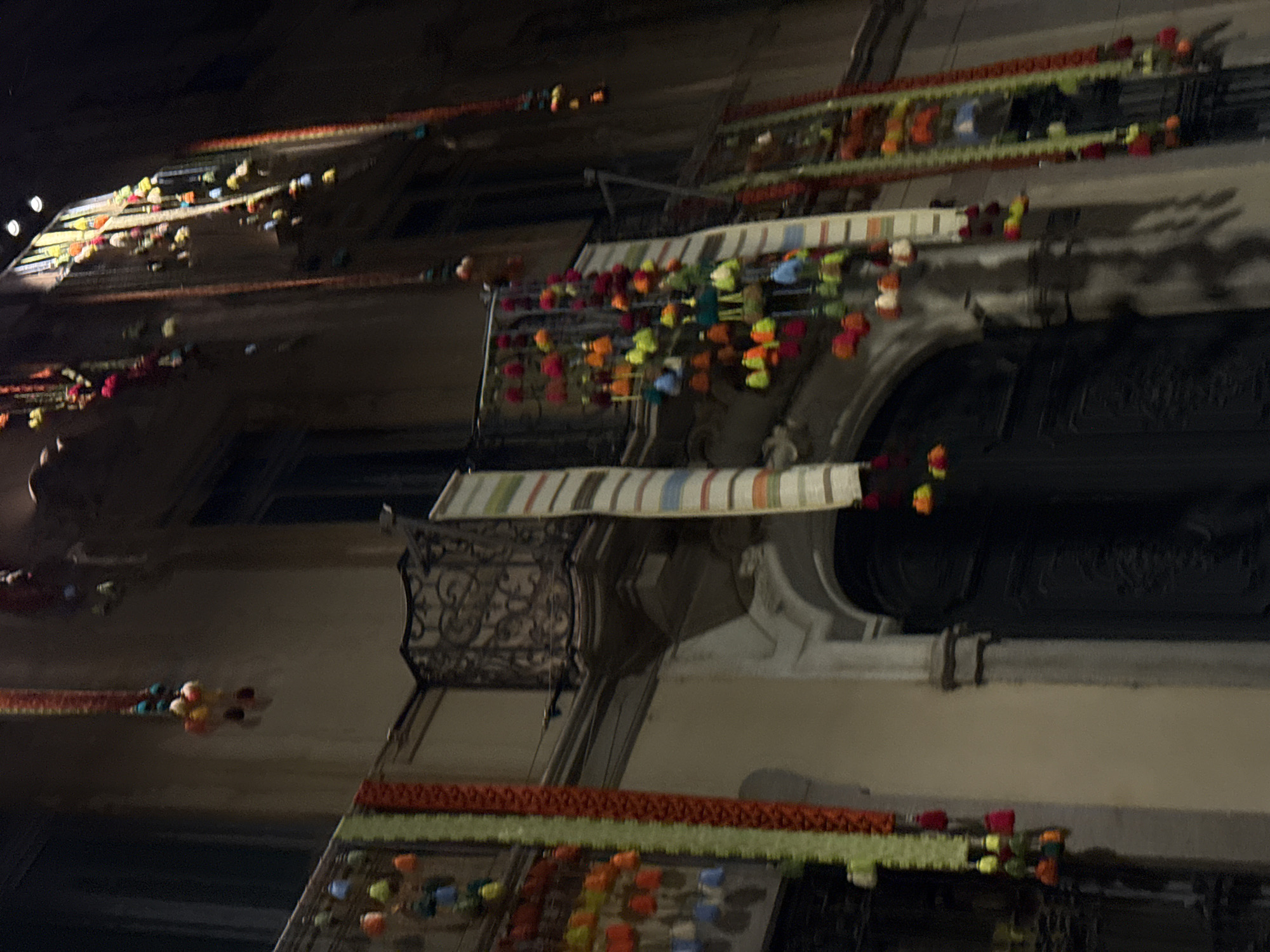 The image displays a nighttime street scene in Milan, Italy, featuring multiple building facades adorned with vibrant decorations, indicative of a festival or celebration. The view is angled upwards, showcasing several balconies on multi-story buildings.
Each balcony is elaborately decorated with long garlands constructed from numerous small, colorful elements resembling pom-poms or fabric flowers. These decorations are predominantly in shades of red, orange, yellow, green, and blue, strung horizontally across the balconies and cascading downwards. The garlands create a rich tapestry of color against the dark facades.
Further enhancing the festive atmosphere, several striped tapestries or rugs are draped over the railings of some balconies. These textiles feature vertical stripes in muted tones such as beige, white, brown, blue, and green. The buildings exhibit classical architectural details, including dark, ornate wrought-iron balcony railings, window frames, and decorative moldings, though these are dimly lit.
The overall scene conveys a sense of festivity and community celebration. The time of day is clearly night, with the colorful decorations appearing illuminated against the dark background. No people are visible, and no discernible text appears in the image.
