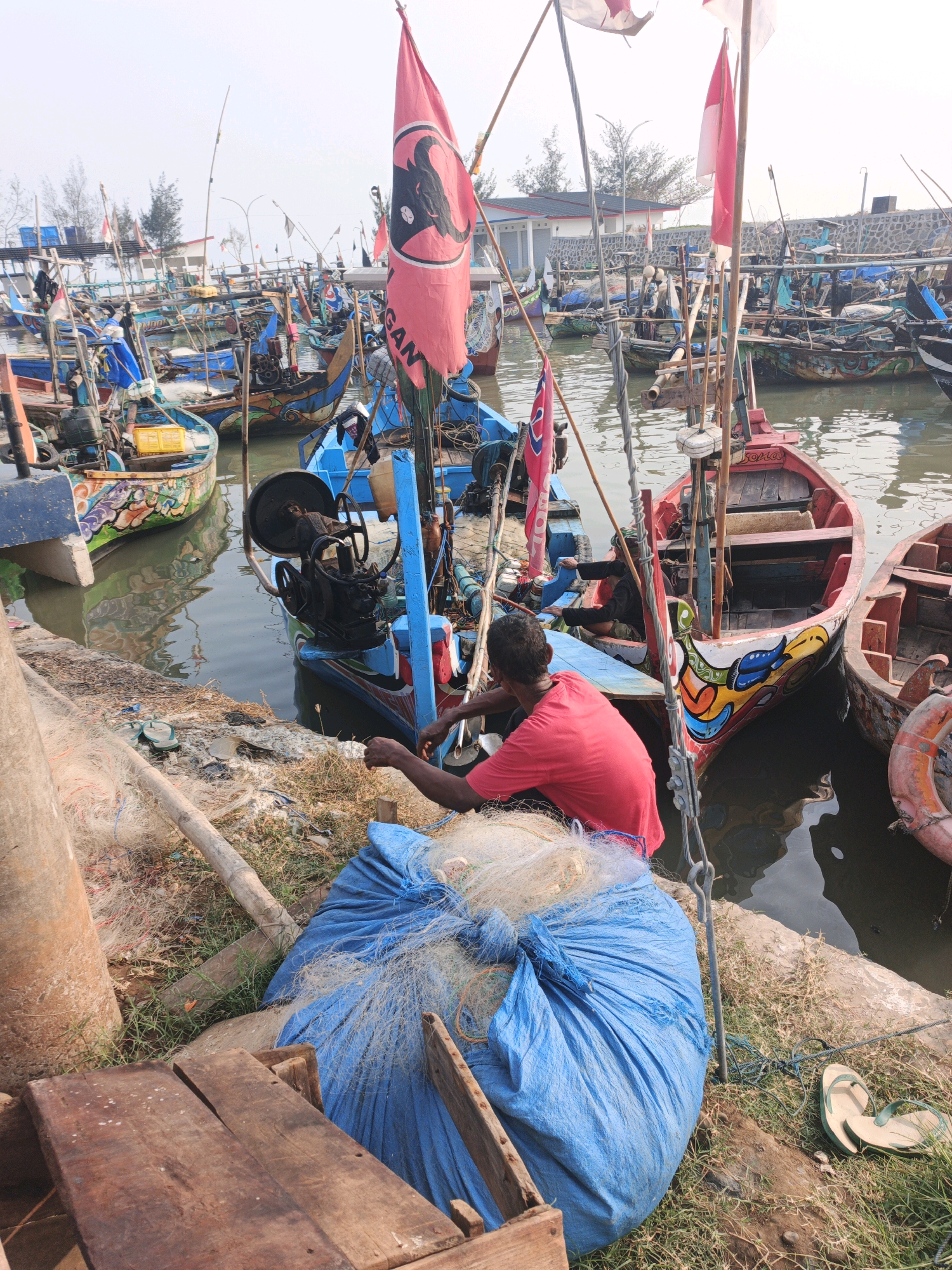 The image depicts a harbor scene in Indonesia, likely a fishing village, with numerous boats docked along a waterfront. In the foreground, a man in a red shirt is seated on the ground, tending to a large blue tarp bag filled with fishing nets. His hands are engaged with the nets, suggesting he is mending or organizing them. Several other fishing boats, characterized by their colorful designs and flags, are visible in the water. Some boats have engines attached. The environment appears to be an outdoor area with dry grass and sand, and a concrete structure or wall along the water's edge. The sky is overcast, indicating a possibly hazy or cloudy day. The overall impression is one of a working fishing harbor.
