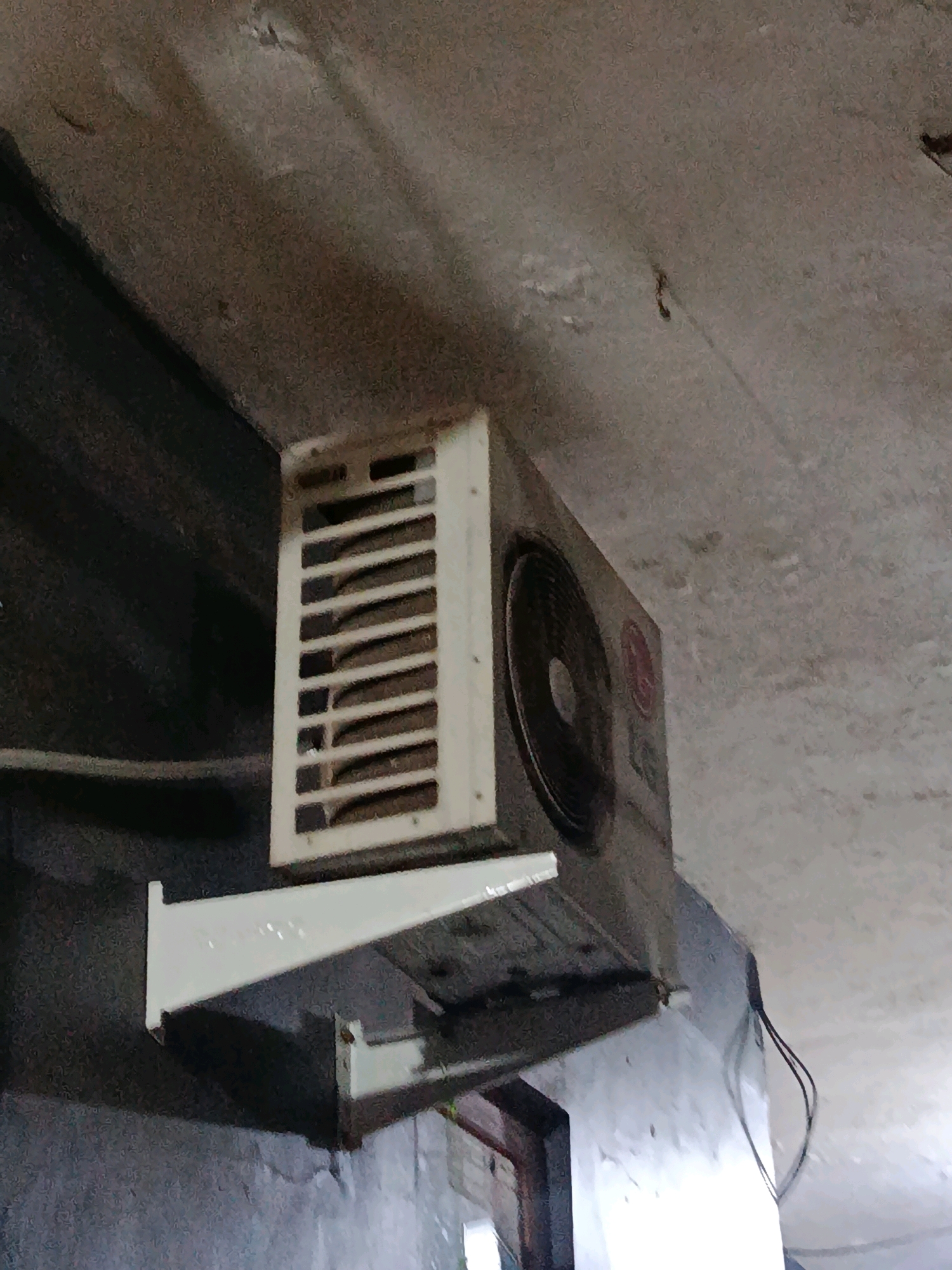 An outdoor air conditioning unit is mounted on a wall with a bracket. The unit is white and has a grille on the front. The fan inside the unit is visible. The wall is dark gray and appears to be made of concrete. The ceiling is also concrete and is textured. There are some wires hanging from the unit. The scene is likely in an outdoor area.