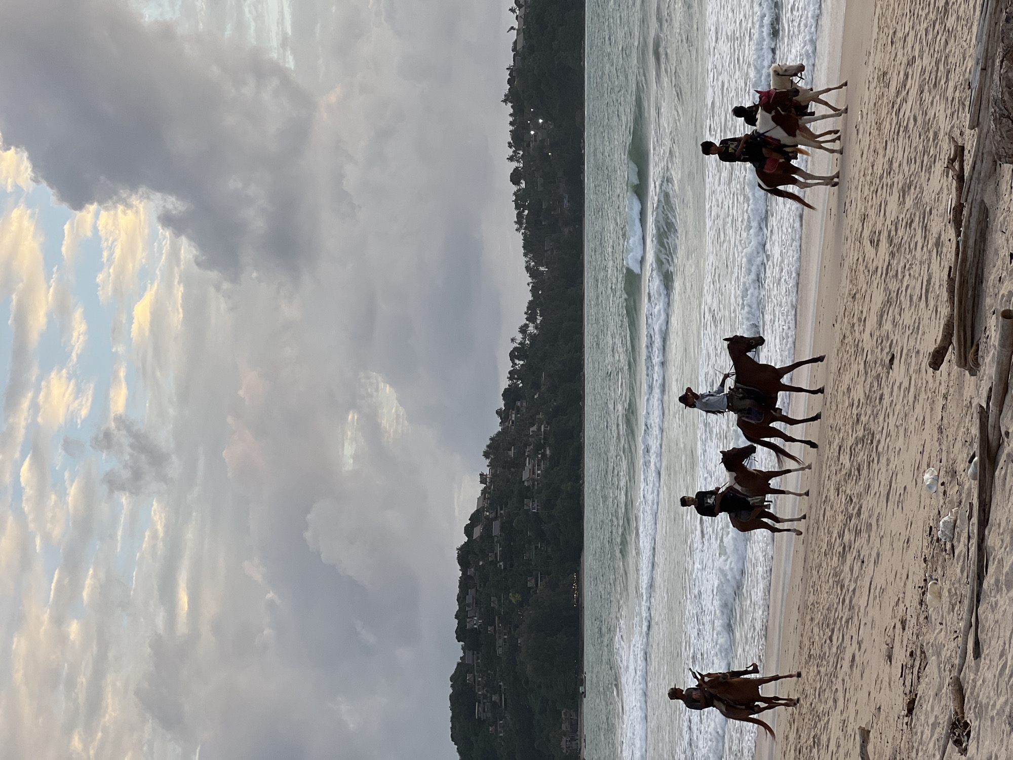The image captures a tranquil scene on a beach in Choeng Thale, Thailand, during what appears to be late afternoon or early evening. The foreground is dominated by a sandy beach littered with driftwood and some discarded plastic bottles. In the middle ground, five people are riding horses along the shoreline. The riders are spaced out, with one rider on the far left, two riders in the center, and two riders on the far right. The horses are of varying colors, mostly brown and white, and are walking calmly along the wet sand where the waves are gently lapping.
The background features the Andaman Sea with moderate waves breaking on the shore, creating white foam. Beyond the sea, a lush green, tree-covered hillside rises steeply, dotted with numerous buildings, suggesting a developed coastal area with residential or resort properties. The sky above is filled with dramatic clouds, a mix of grey and white, with hints of soft yellow and pink light breaking through, indicating the approach of sunset or a cloudy evening. The overall atmosphere is serene and picturesque, highlighting a popular tourist activity in the region.