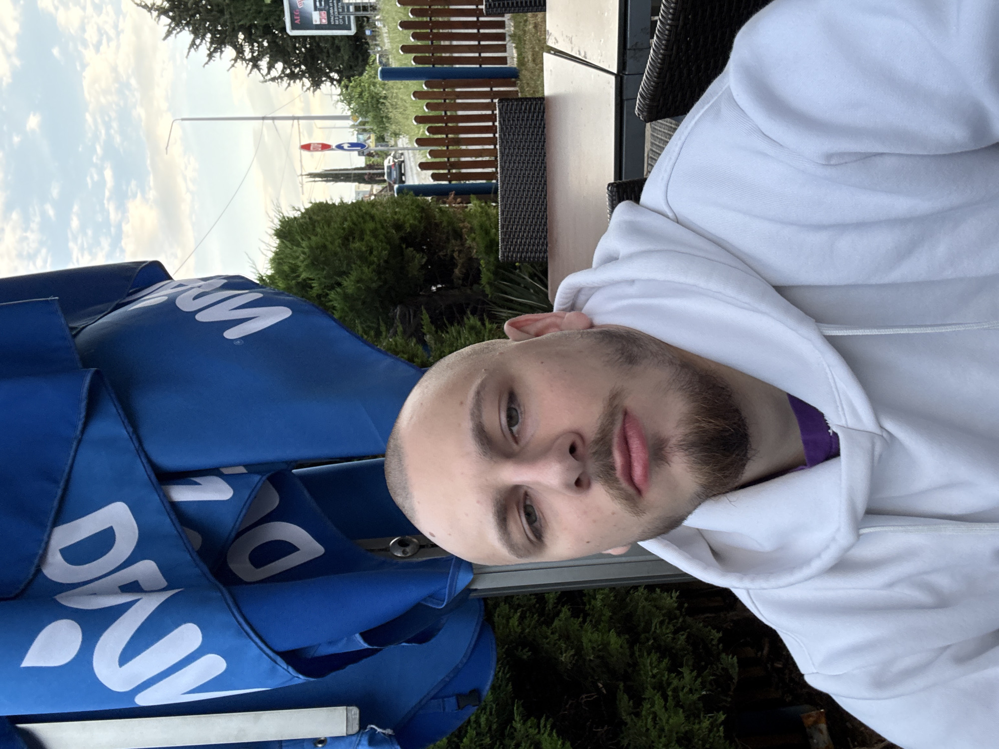 A person with short hair and a beard is visible in the foreground, wearing a white hoodie. A partially visible blue parasol with white text is adjacent to the person. In the background, there is a street scene with greenery, a wooden fence, street signs, parked cars, and buildings, suggesting an outdoor café setting. The location is inferred to be in the city of Stara Zagora, Bulgaria.