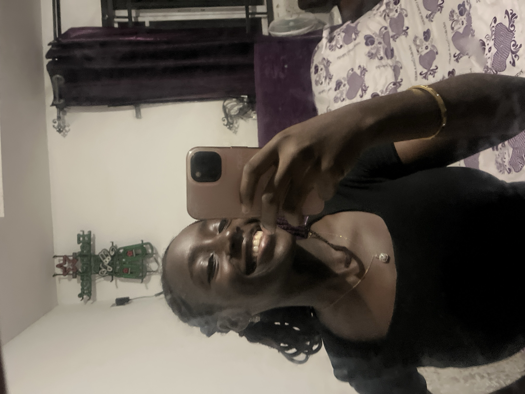 A young Black woman is taking a mirror selfie in what appears to be a bedroom in Abuja, Nigeria. The woman is smiling broadly, showing her teeth, and her eyes are closed. She is wearing a black top and a gold necklace with a pendant. A gold bracelet is visible on her right wrist. Her hair is braided and pulled back. In the background, on the wall to the left, is a green decorative item, possibly a Christmas ornament, with red accents and a star at the top. To the right, dark purple curtains hang from a curtain rod. Behind the woman, a bed with white bedding featuring a pattern of purple hearts and what appear to be teddy bears is visible. A fan and part of a headboard are also present in the background. The lighting suggests an indoor, possibly evening setting.