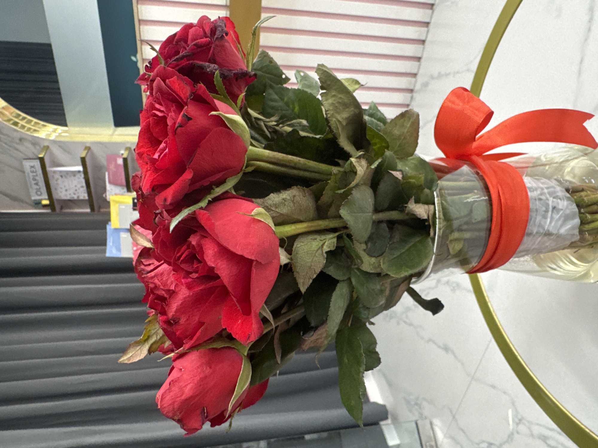 The image depicts a bouquet of red roses wrapped in clear plastic and tied with a red ribbon. The bouquet is positioned in front of a gray and white marble background. The background contains some furniture and beauty salon signage reading "Olaplex." The location is North Kuta, Indonesia.