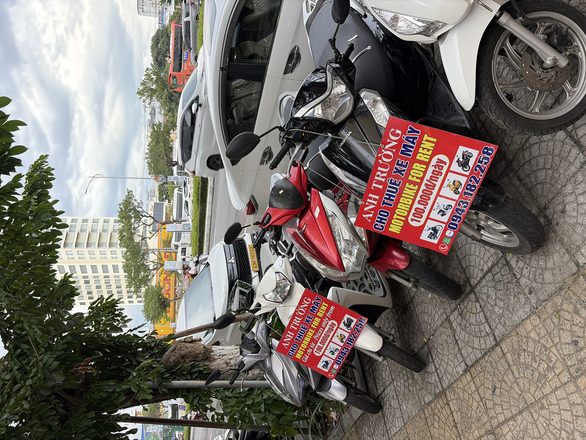 A street scene in Da Nang, Vietnam, features several parked motorbikes for rent, indicated by red signs with Vietnamese text and English translations. The signs display a phone number, "0943.182.258," and prices, "100.000đ/ngày" (100,000 VND/day), along with icons representing different motorbike models. The motorbikes are parked on a paved sidewalk next to a street with moving traffic. In the background, a white car and a tall, modern building are visible. The sky is overcast.