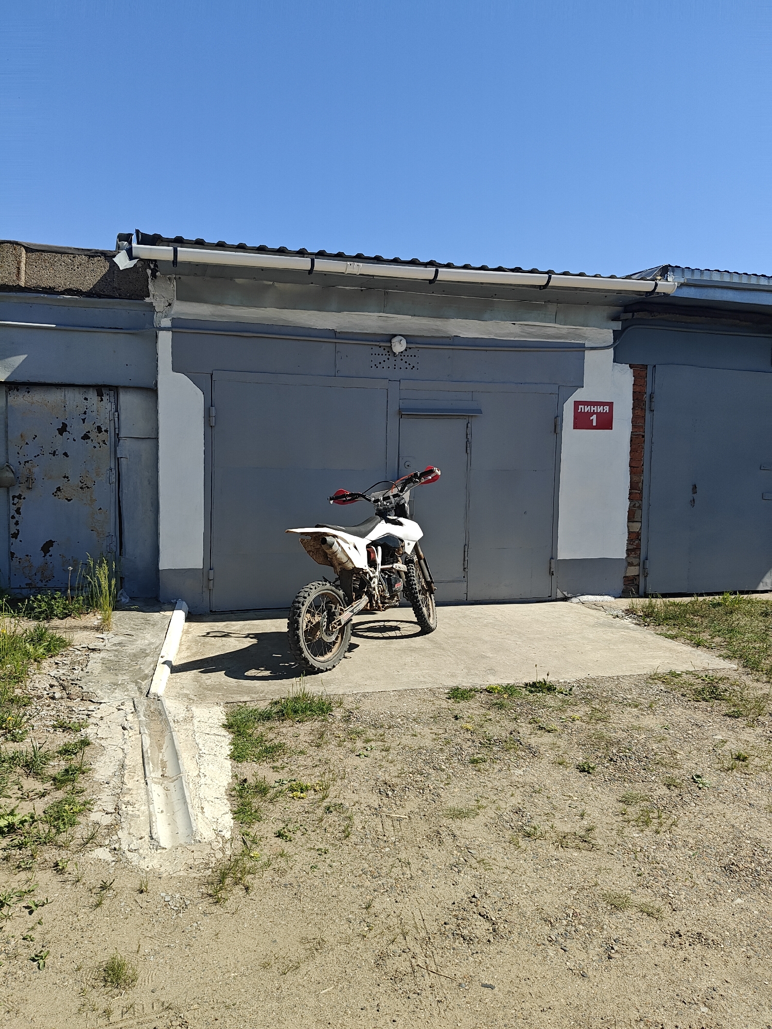 A dirt bike is parked in front of a row of gray garage doors in Chaikovsky, Russia. The doors are numbered in red as "ЛИНИЯ 1." The parking surface is sandy with grass and weeds growing around the edges. The sky is clear and blue. No people are visible.