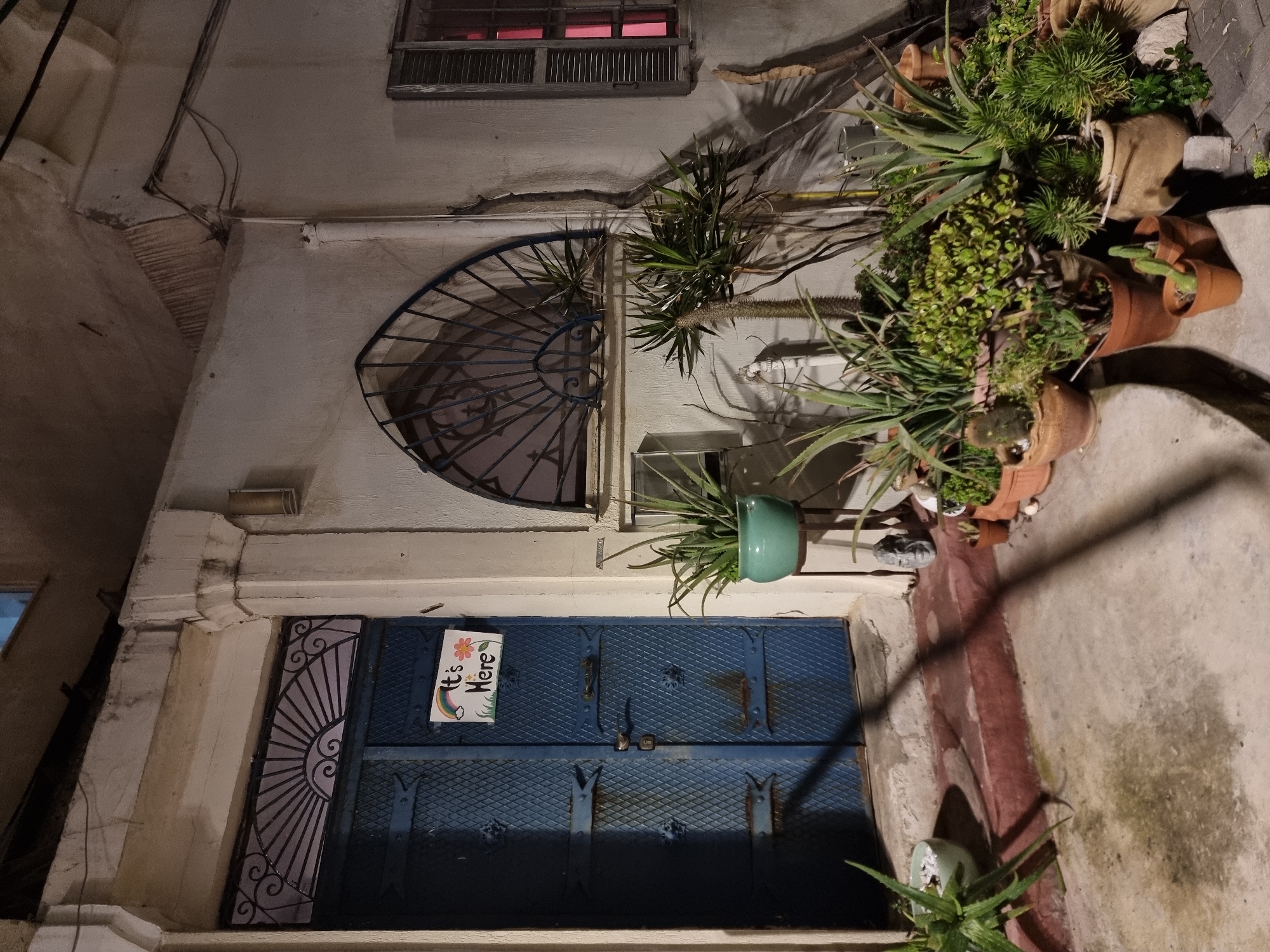 The image captures a doorway in Tel-Aviv, Israel. The door is blue with a metal grate pattern and a smaller decorative metal panel on the left side. A sign is attached to the door, reading "It's Here" with drawings of a rainbow, flower, and stars. Above the door is a window with a decorative blue metal grate. Above that window is a small, shuttered window with red light visible inside. Various potted plants surround the doorway on the right, including succulents and other greenery. The building's facade is primarily white and appears to be aged. The scene is dimly lit.