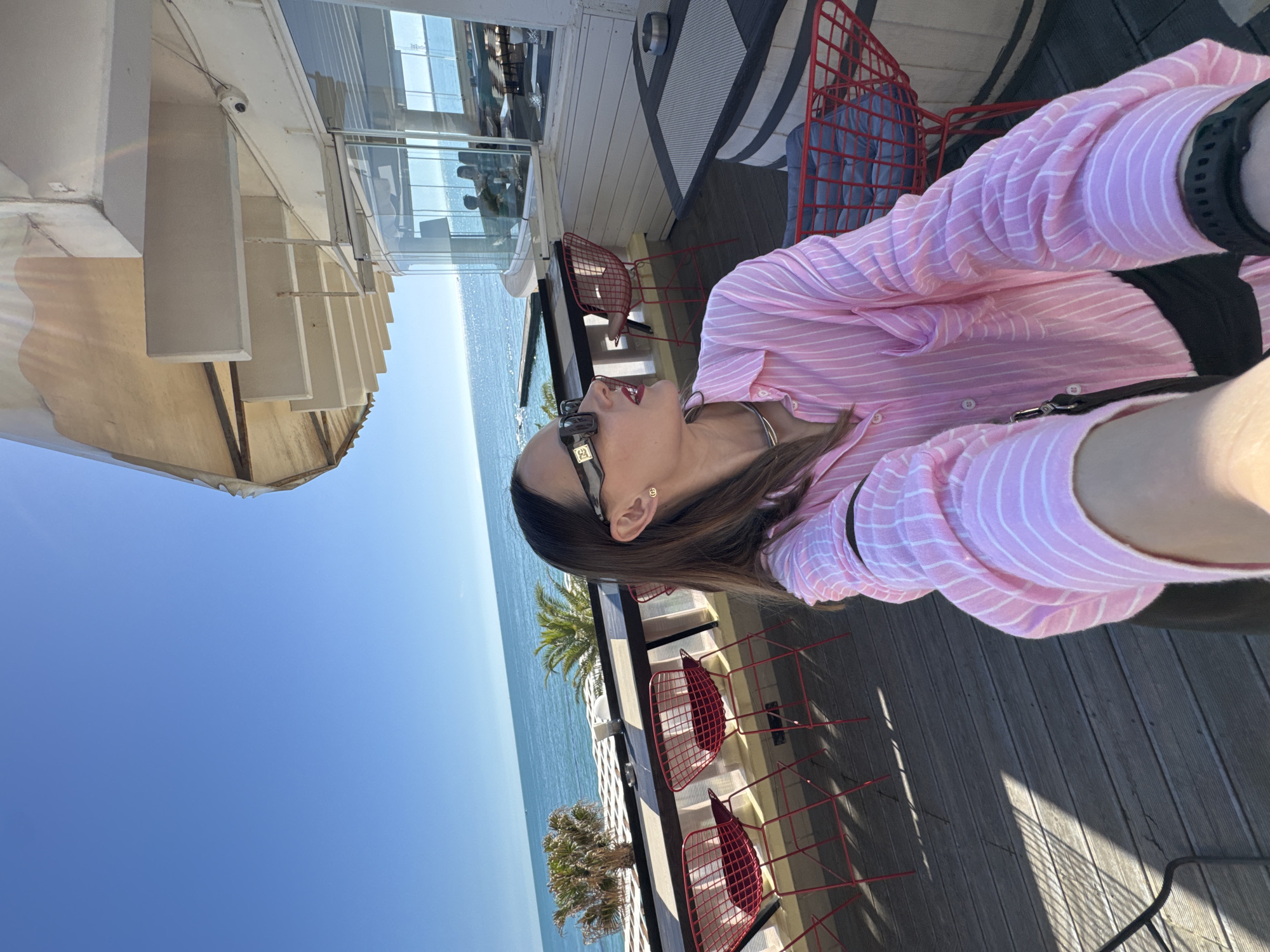 A bright and sunny outdoor scene captures a woman relaxing on a balcony or terrace overlooking the sea in Sochi, Russia. The image is oriented vertically, showing the woman in the foreground on the right side of the frame, facing left and looking upwards. She is wearing a light pink and white striped long-sleeved button-up shirt, stylish black sunglasses with gold details, and a black watch on her left wrist. Her brown hair is visible around her face and shoulder.
The setting is a wooden deck, likely part of a cafe or restaurant, furnished with several modern red wire-frame chairs, some with blue cushions. A black and white striped table surface is also partially visible. Beyond the seating area, a vast expanse of deep blue sea stretches towards the horizon, meeting a clear, light blue sky. Lush green palm trees can be seen growing below the terrace, enhancing the coastal ambiance.
The architecture includes a white building with large, reflective glass windows, which mirror the sky and sea. Strong sunlight casts distinct shadows on the wooden decking, indicating that it is midday or early afternoon. A small, white security camera is mounted on the underside of a white overhead structure. The overall impression is one of leisure and enjoyment of a beautiful, sunny day by the sea.