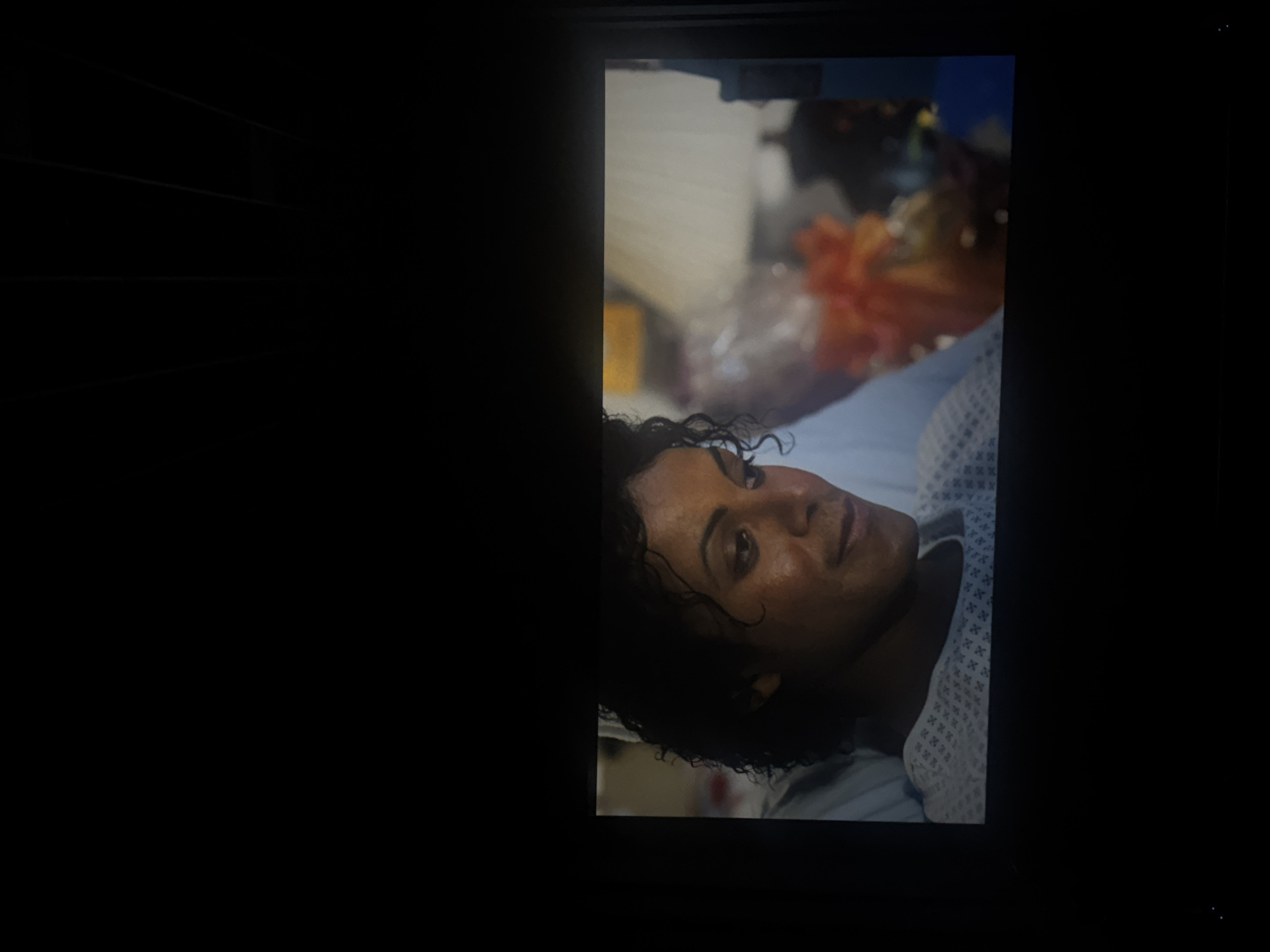 A person with dark hair and skin is lying on a light blue and white patterned hospital bed. The person is looking towards the viewer. Behind the person is what appears to be hospital equipment and medical supplies. The location is not discernible from the image.