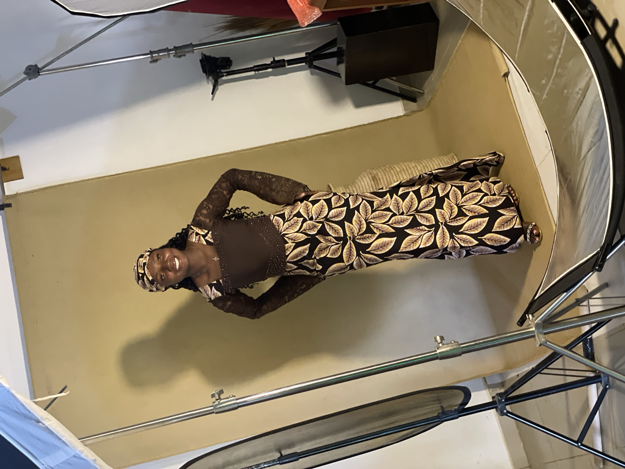In Durumi, Nigeria, a young Black woman is posing for a photo shoot in a studio setting. She is wearing a long, brown dress with a patterned skirt featuring large, light-colored leaves on a dark background. The top of the dress is brown with lace sleeves, and she has a matching headscarf. She is smiling and has her hands on her hips, looking towards the camera. Behind her, a large, light-colored backdrop is set up, and studio lighting equipment is visible, including stands and diffusers. A tripod with a camera is also present in the background, along with a dark, rectangular pedestal. The floor is covered with a light-colored mat.