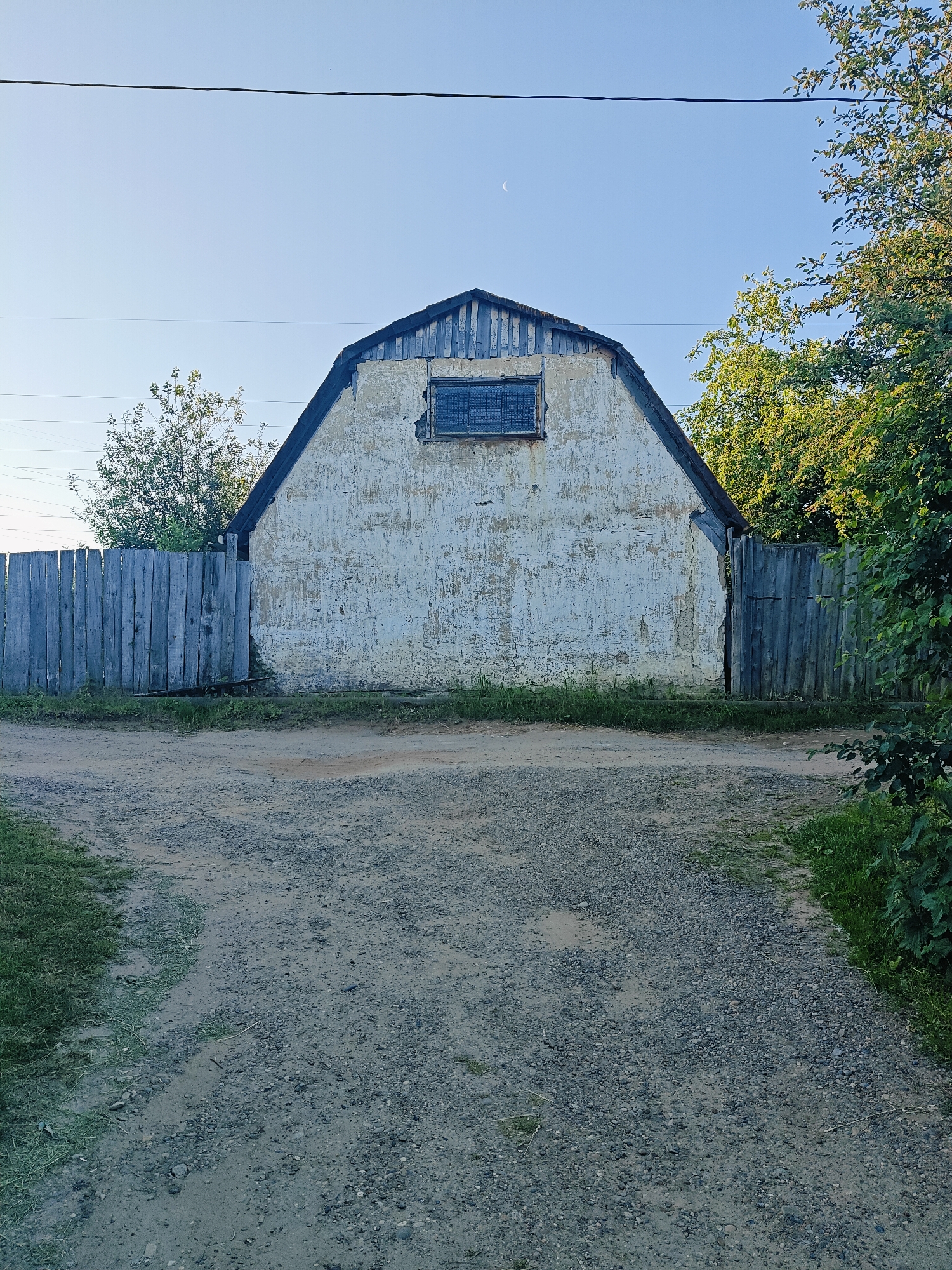 A detached, gambrel-roofed building with peeling white stucco exterior is situated behind a weathered, grey wooden fence. The building has a small, barred window near the peak of the roof. Tall green trees and foliage surround the property on the right and behind the fence. A gravel path leads towards the building, splitting into two distinct tracks. Overhead, a thin black wire stretches across the frame, and a sliver of the moon is visible in the clear blue sky. The scene is consistent with a rural or semi-rural area within the city of Chaikovsky, Russia, possibly a private residence or an outbuilding.