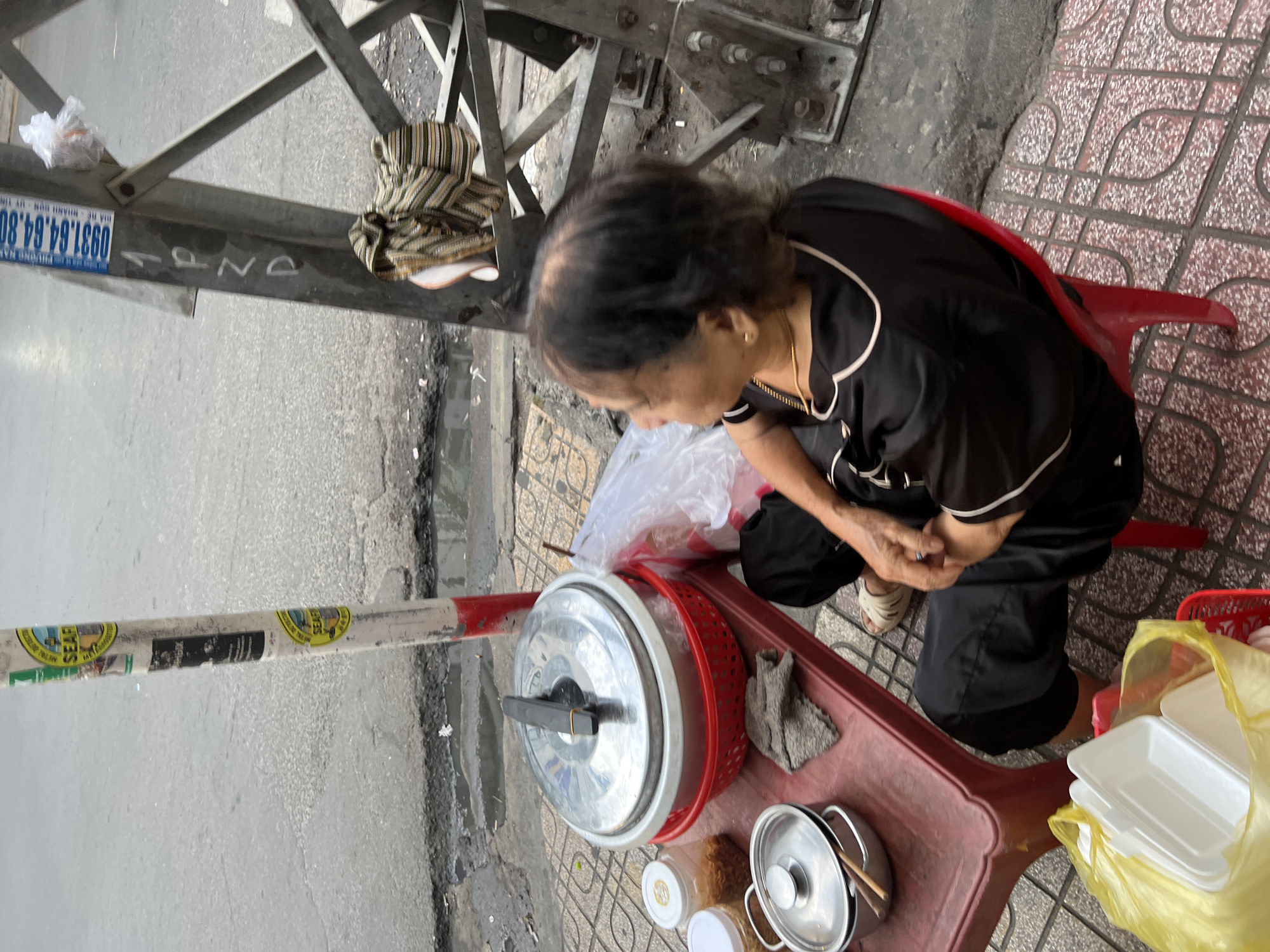 An elderly woman wearing a black top and black pants sits on a red plastic chair on a sidewalk in Nha Trang, Vietnam. She is seated next to a small red plastic table. On the table are several food containers, including a large silver pot with a lid, a red plastic basket containing another silver pot, and two glass jars with lids. To the left of the table, a yellow plastic bag holds white styrofoam food containers. A metal pole with various stickers and markings stands behind the table and to the right of the woman. A section of a power line tower is visible to the right of the pole. The sidewalk is made of patterned tiles and abuts a street with visible water puddles and cracks. A rolled-up striped fabric is draped over a part of the power line tower. The woman appears to be waiting or serving food.