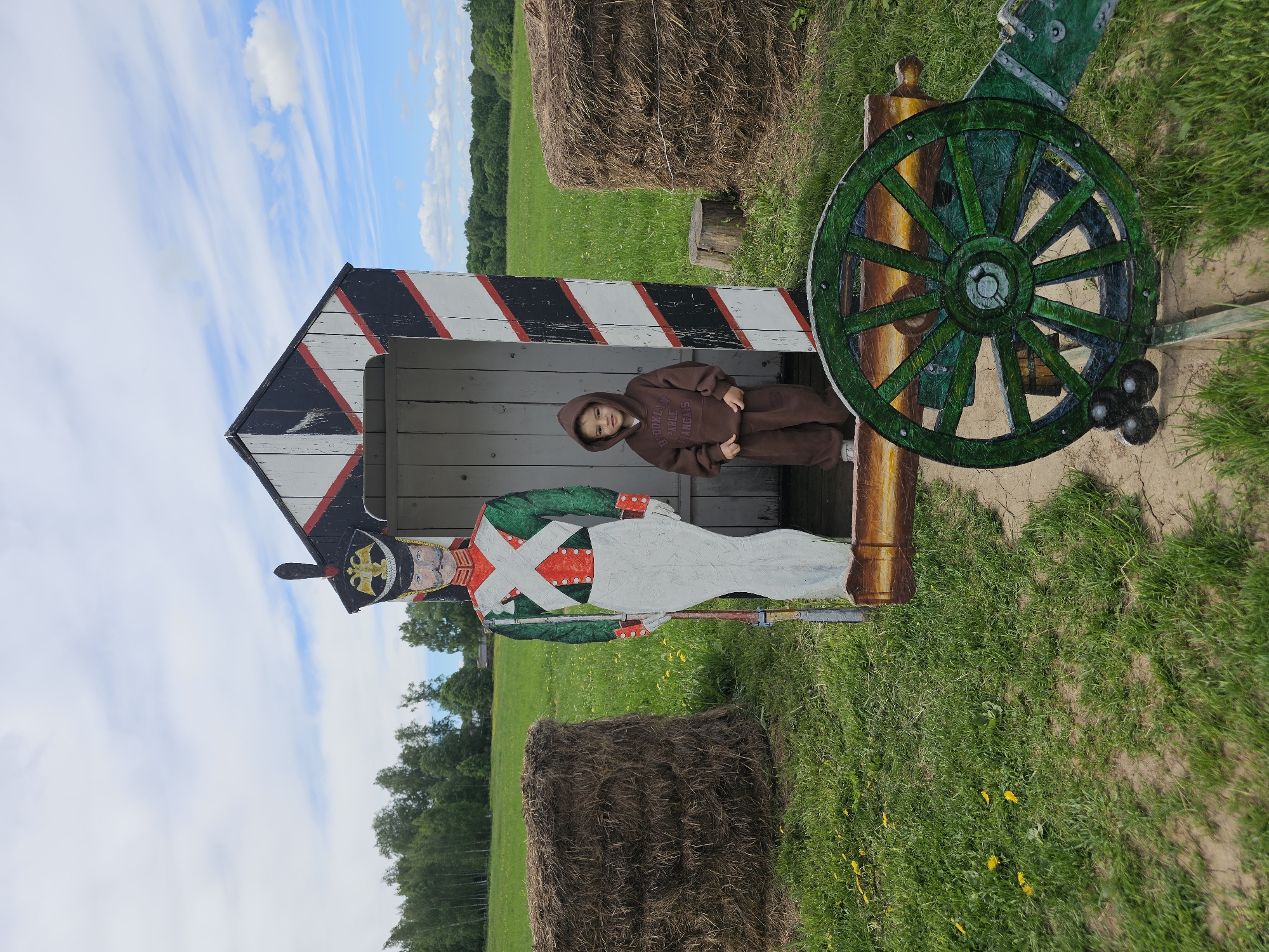 A child in a brown hooded sweatshirt is seated inside a black and white diagonally striped sentry box. A painted wooden cutout of a soldier stands to the left of the child, and a wooden cannon with a green wheel is to the right. The setting is a grassy field, possibly in the area of Borodino, Russia, considering the Napoleonic-era soldier representation. Hay bales are in the background. The sky is partly cloudy.