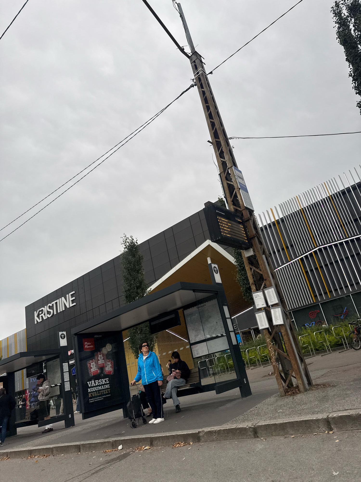 The image depicts the exterior of a Kristiin Center shopping complex located in Tallinn, Estonia. A bus stop with a shelter is visible in the foreground, with several individuals waiting. One woman in a blue jacket stands with a rolling suitcase, while another person sits on a bench within the shelter, looking at their phone. A third person, wearing a light-colored jacket and dark pants, is also seated on the bench. In the background, the modern architecture of the Kristiin Center is apparent, characterized by dark cladding and a prominent white "KRISTIINE" sign. The sky is overcast, and electrical wires are strung across the frame, supported by a weathered metal pole near the bus stop. Adjacent to the shopping center is an area with numerous bicycle racks.