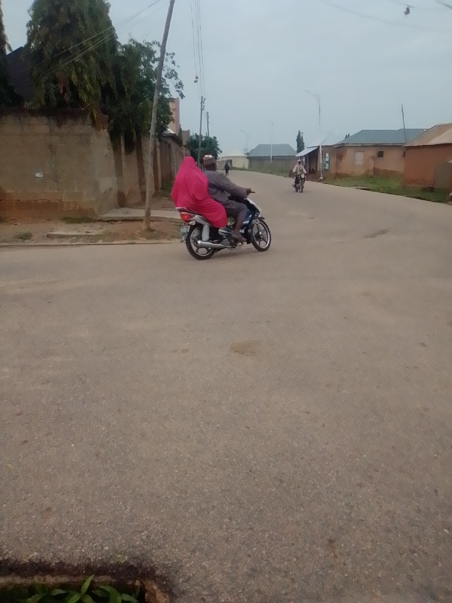 In Gombe, Nigeria, two individuals are riding on a motorcycle on an asphalt road. The person in the back is wearing a bright pink garment that covers them from head to toe, with only their face partially visible. The person in front is wearing a cap and a gray garment. The motorcycle is a light color. The road is lined with buildings and trees. The sky is overcast. The scene appears to be a typical street in a residential area.