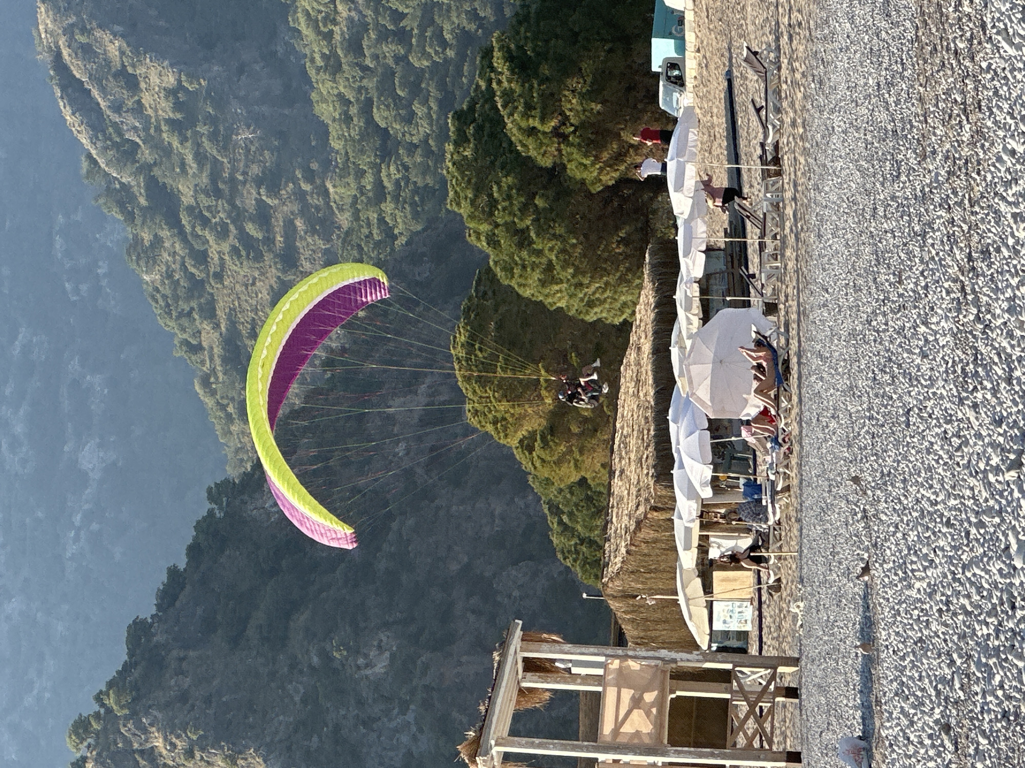 A paraglider with a vibrant green and purple canopy descends over a pebbly beach and forested mountains in Fethiye, Turkey. Two individuals are harnessed beneath the paraglider, actively soaring through the air.
In the foreground, a sun-drenched beach stretches across the frame, characterized by numerous white beach umbrellas and an assortment of lounge chairs. Several people are visible relaxing on the beach, some seated under the umbrellas, enjoying the sunny weather. A rustic, wooden beach structure with a thatched roof is positioned on the left side of the beach area. Behind the beach, a dense line of green trees separates the shore from the towering, verdant mountains that dominate the background. These mountains appear steep and are covered in thick foliage.
Vehicles, including a white van and a light blue truck, are parked behind the beach, partially obscured by the trees. The bright sunlight suggests it is either late morning or afternoon, casting some shadows. The sky is clear. On a small sign or menu near the beach structure, some indistinct text is visible, likely listing offerings for beachgoers, though it is illegible. The scene captures a popular tourist destination, combining adventurous paragliding with relaxing beach activities.