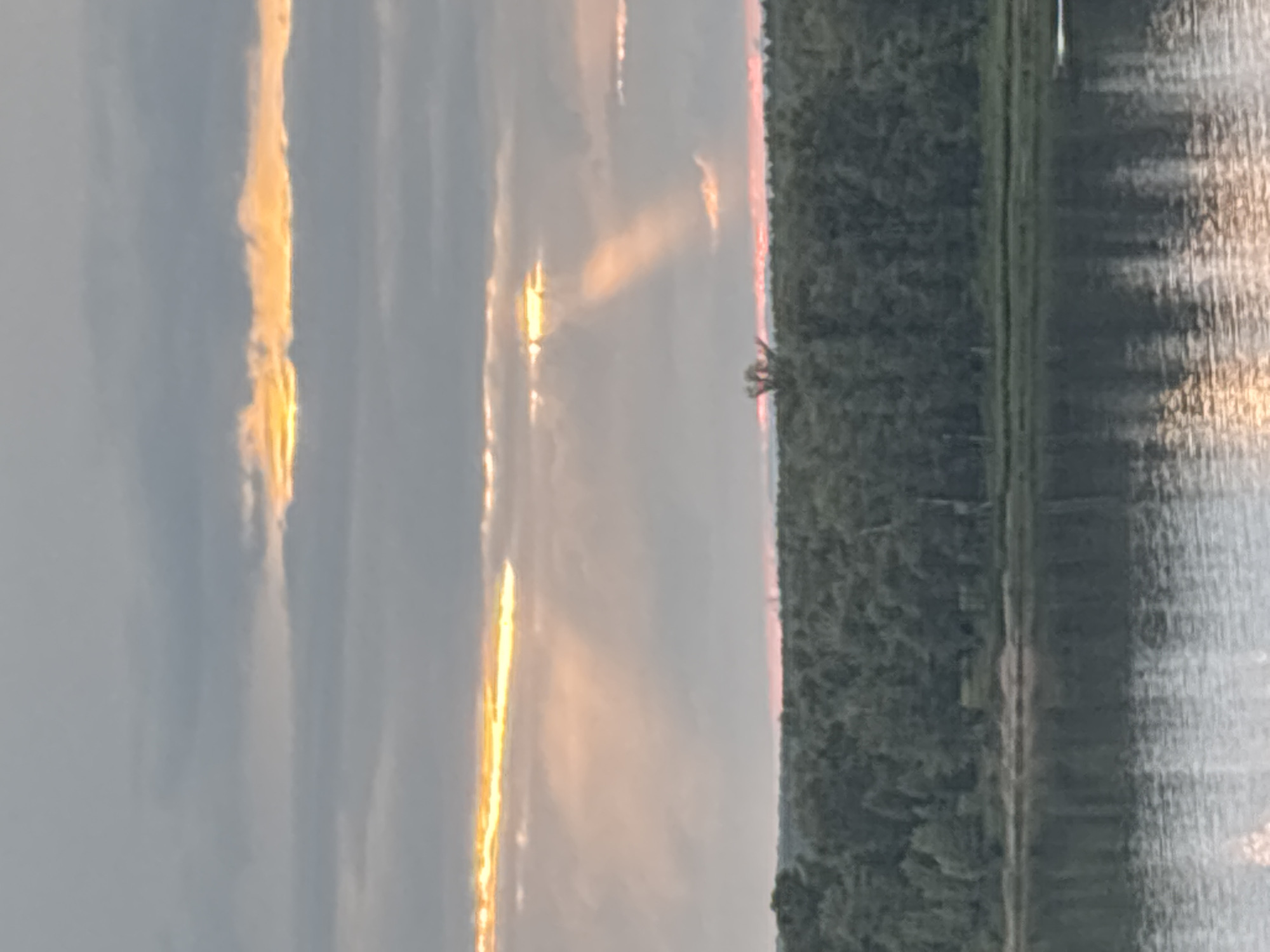 This media file captures a scenic view of a sunset over a body of water, likely a river, in Olkhovka, Russia. The image is oriented vertically, with the sky and clouds dominating the left side and the water and treeline on the right.
The sky is filled with dramatic cloud formations, illuminated by the setting sun. Streaks of golden light pierce through the grey clouds, casting warm hues and creating a dynamic interplay of light and shadow. A soft pinkish glow is visible near the horizon, indicating the approaching twilight.
On the right side of the image, a dense treeline forms a dark silhouette against the colorful sky. The trees appear lush and green, suggesting a summer or early autumn setting. The water's surface reflects the sky's light, with shimmering patterns of gold and grey creating a textured effect. The water appears calm, with only gentle ripples disturbing its surface.
A single, small tree stands out against the treeline, providing a point of interest in the otherwise unbroken expanse of foliage. The overall mood of the image is one of tranquility and natural beauty, enhanced by the warm glow of the sunset. The absence of any people or man-made objects emphasizes the serene and untouched quality of the natural landscape. The time of day is clearly sunset, and the weather appears clear, allowing for the vibrant display of colors in the sky. There is no visible text in the image.