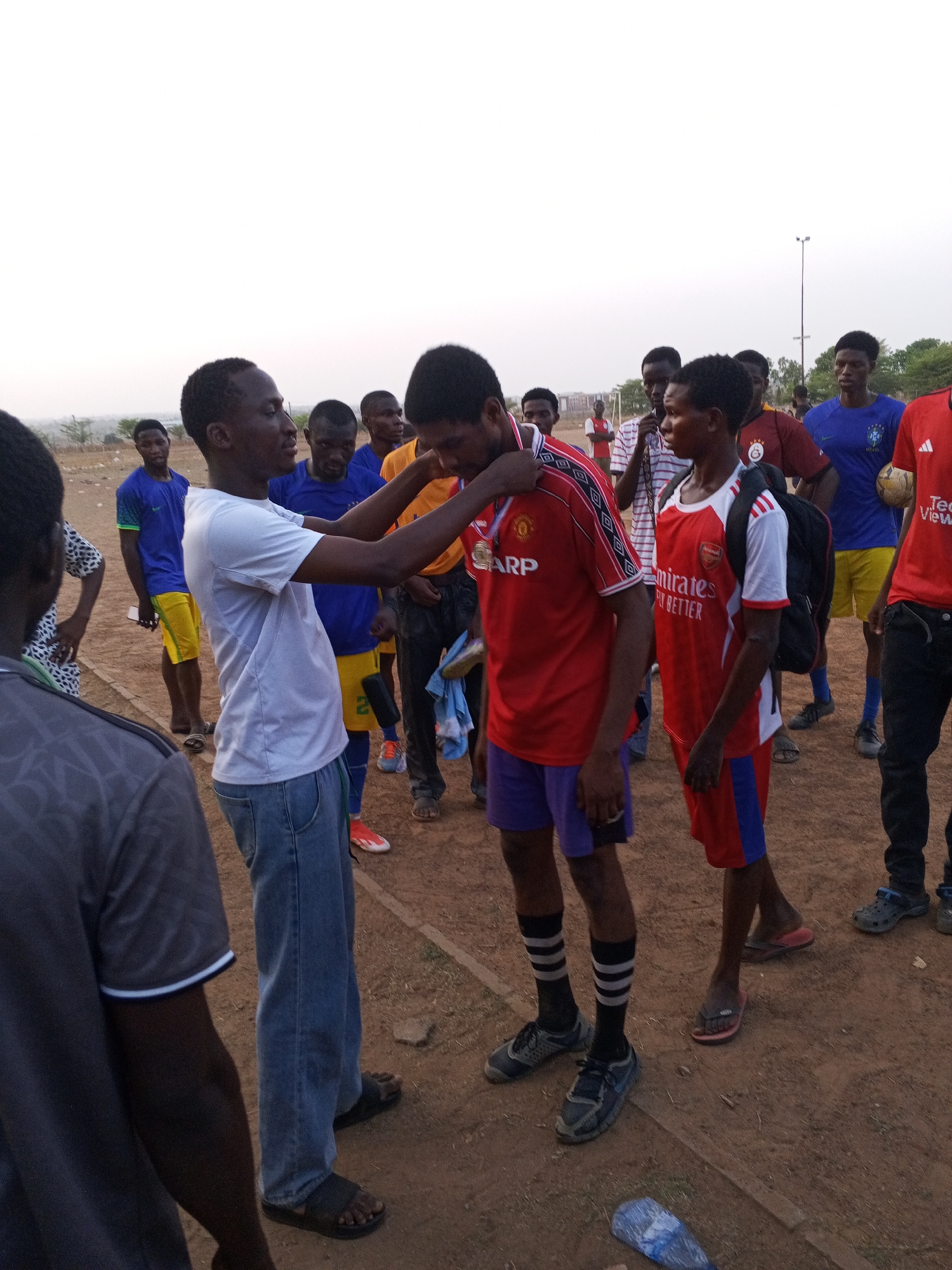 In Minna, Nigeria, a group of young men stands on a dirt field. One man in a white t-shirt and blue jeans is placing a medal around the neck of another man wearing a red Manchester United soccer jersey and purple shorts. The surrounding men wear a variety of soccer jerseys and casual attire. The environment includes a soccer field with a light pole and scattered trees.