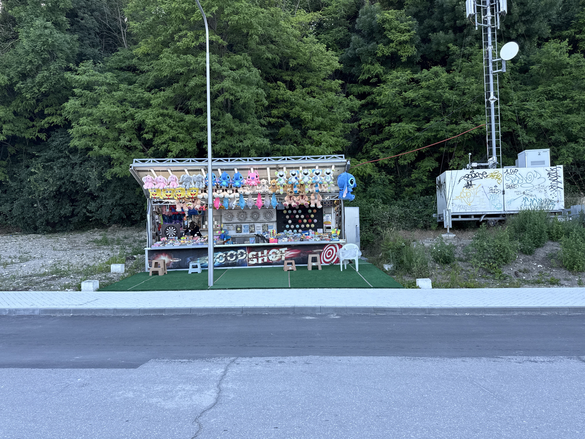 A fairground stall is set up on a grassy area, with a road in the foreground and a dense, green forest behind it. The stall is a long, narrow structure with a roof and an open front. It is stocked with rows of stuffed animals, primarily cartoon characters in various colors and sizes, hanging from the roof. Below the stuffed animals, there are game booths with targets, such as a dartboard and circular targets. The stall is illuminated by a street lamp. To the right of the stall, a metal cell tower stands tall, with a white rectangular structure at its base covered in graffiti. The environment suggests a recreational area or roadside attraction adjacent to a wooded hillside.
