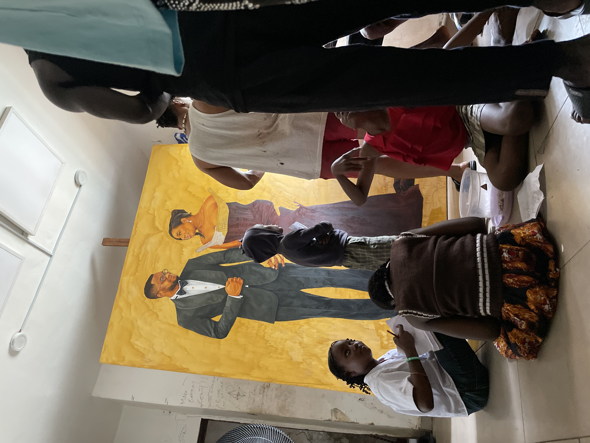 In Ado, Nigeria, within a room with a light-colored ceiling and walls, a large, unfinished painting is displayed on an easel. The painting depicts a man in a tuxedo and a woman in a formal gown, both with serious expressions, against a vibrant yellow background. Several individuals are present, some actively engaged with the artwork and others observing. A young girl in a white top and dark pants sits on the floor, holding a pencil and paper, seemingly drawing or writing. To her right, another person, seen from the back, is wearing a brown vest over a striped shirt and a patterned skirt, also seated on the floor. A child in a dark hoodie stands near the painting. Several adults are partially visible, one in a white tank top and red shorts, and another in black pants. The floor is tiled, and various objects are scattered around, including a plastic container. The overall atmosphere suggests an art studio or a space dedicated to creative activity, possibly a community art project.