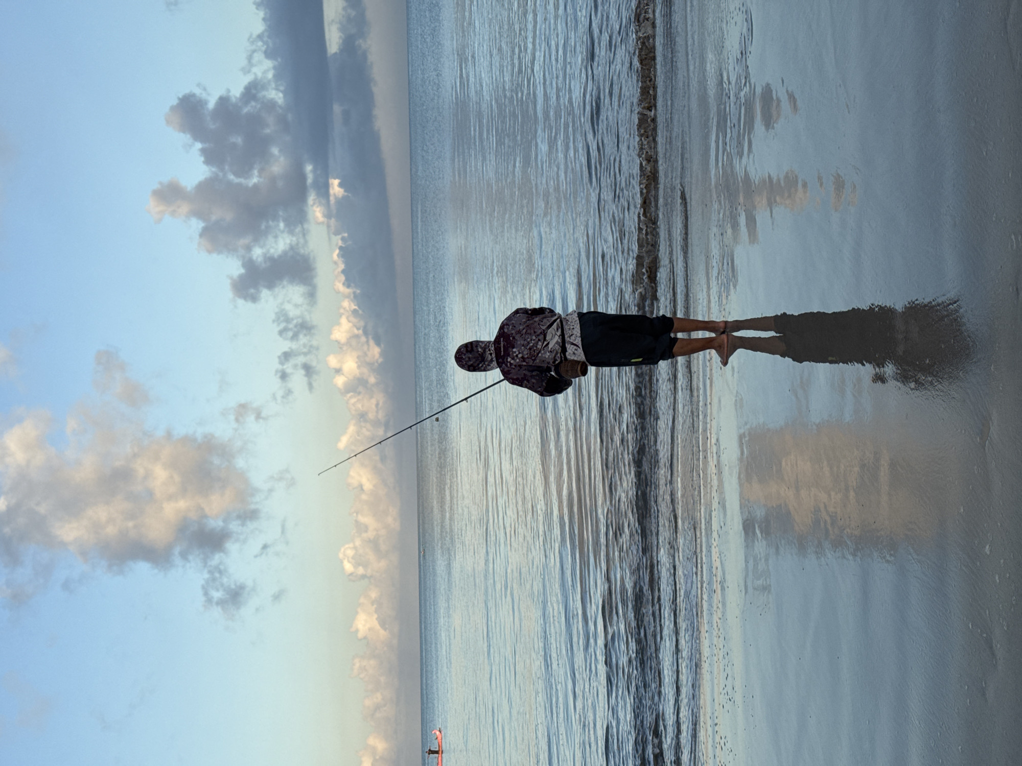 A person is fishing on the beach in Kuta, Indonesia. The individual, dressed in shorts and a hooded top, stands at the edge of the water, casting a fishing rod out to sea. A clear reflection of the person and their shadow stretches out on the wet sand. In the distance, calm water meets the horizon under a sky of blue and cloud formations. A small boat can be seen floating on the horizon.
