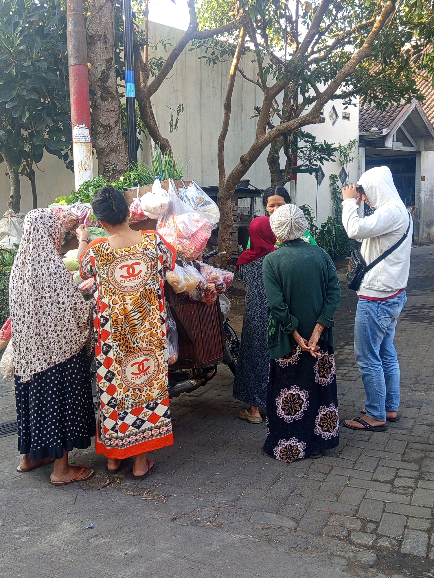 In Singosari, Indonesia, on a paved street lined with trees and a building with white walls and a tiled roof, four individuals are gathered around a mobile fruit and vegetable cart. The cart is laden with produce in plastic bags. One woman, wearing a patterned headscarf and a polka dot skirt, stands on the left. Next to her, a woman in a vibrant, patterned dress with a Chanel logo and an animal print motif is interacting with the cart. Another woman, with a red headscarf, stands near the cart. On the right, a person wearing a white hoodie and blue jeans is holding up a phone, appearing to take a photo. The scene suggests a moment of commerce or social interaction on a bright, sunny day.