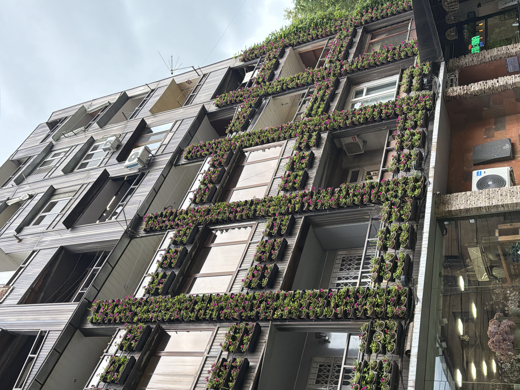 Here is a description of the image:
The image captures a low-angle view of a building adorned with a green facade. The architecture is characterized by geometric, angular elements, with wooden-style panels creating a striped effect. Rows of potted plants, lush with greenery and small pink flowers, accentuate the horizontal lines of the building, creating a vertical garden effect between each window and architectural line.
Some windows appear reflective, while others are more transparent, revealing interior details like white bars. Air conditioning units are visible on some floors. A tall antenna is visible atop the building. The adjacent building to the right is also visible, including air conditioning units and a commercial sign. The sky is overcast. Based on the details of the foliage and architecture, it is likely this photo was taken in Yerevan, Armenia.