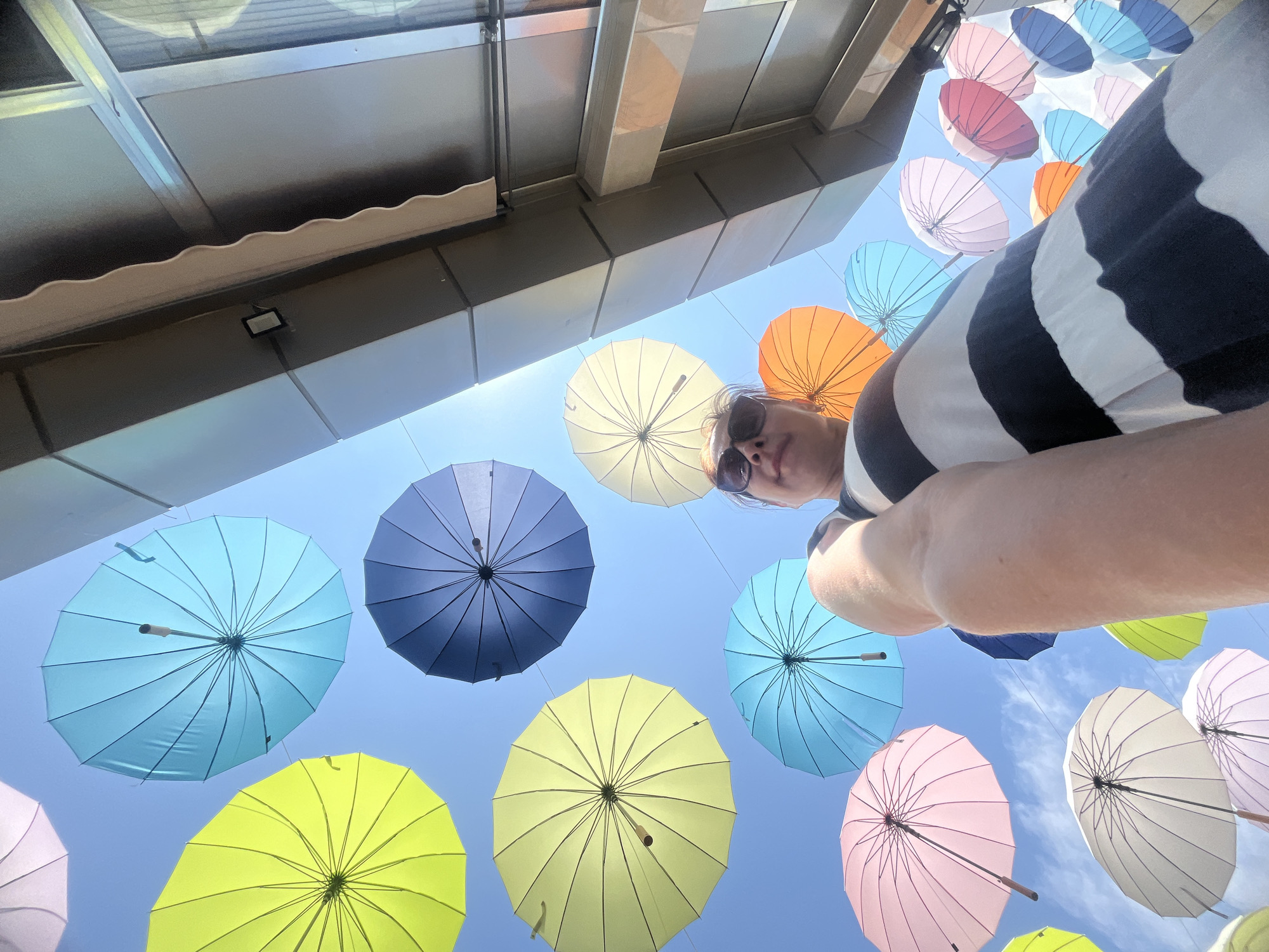 The image depicts a low-angle perspective of a woman in a black and white striped dress, looking up at a vibrant display of colorful umbrellas suspended against a clear blue sky. The umbrellas, in shades of yellow, blue, pink, orange, and white, are arranged in a seemingly random pattern. The woman is wearing sunglasses and her arm is extended forward, possibly holding the camera. In the background, the facade of a modern building, characterized by its metallic panels and glass windows, is visible on the right side of the frame. The lighting suggests a sunny day. This installation of umbrellas is a known sight in the city of Sochi, Russia.
