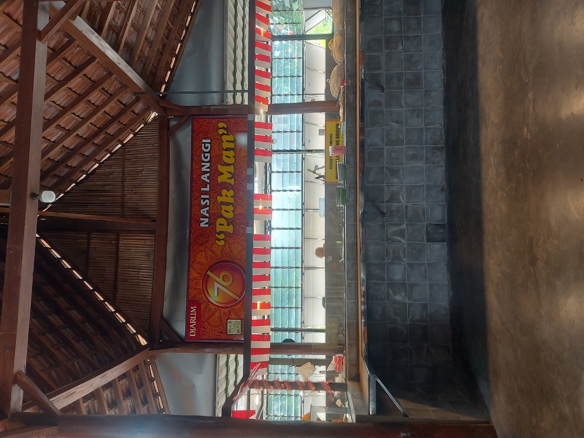 The image shows the interior of an open-air food stall or restaurant, likely in Kricak, Indonesia, characterized by its traditional wooden-beam roofing and bamboo wall accents. A prominent banner hangs above the counter, displaying "DIARUM" in red lettering, followed by "NASI LANGGI 'Pak Man'" in orange and yellow, with a large '76' emblem in the center. Red and white striped fabric is draped across the front of the counter area. The counter itself is tiled with dark grey squares and appears to be empty, save for a few stacks of plates and some food containers visible in the background. Behind the counter, large windows with horizontal louvers offer a glimpse of an outdoor area with greenery. A single person, a woman with blonde hair, is partially visible in the background, seated behind the counter. The overall impression is that of a food establishment preparing to open or recently closed.
