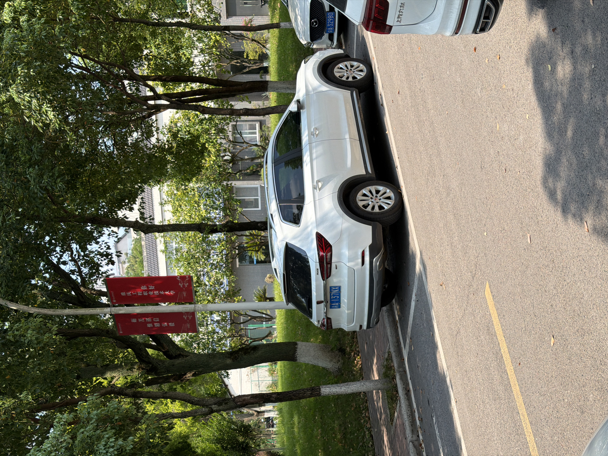 The image depicts a street scene in Jiangjin, China, on a sunny day. A white SUV is parked on the right side of the asphalt road, next to a curb and a grassy area. The SUV has a license plate that reads "A 137NX". A portion of another white car is visible on the far right of the frame. In the background, there are trees with green foliage and a building. A red banner with Chinese characters hangs on a lamppost between the trees and the building. The banner appears to be advertising a university. The overall environment suggests a park-like setting within an urban area, possibly a campus or a residential neighborhood with ample greenery.