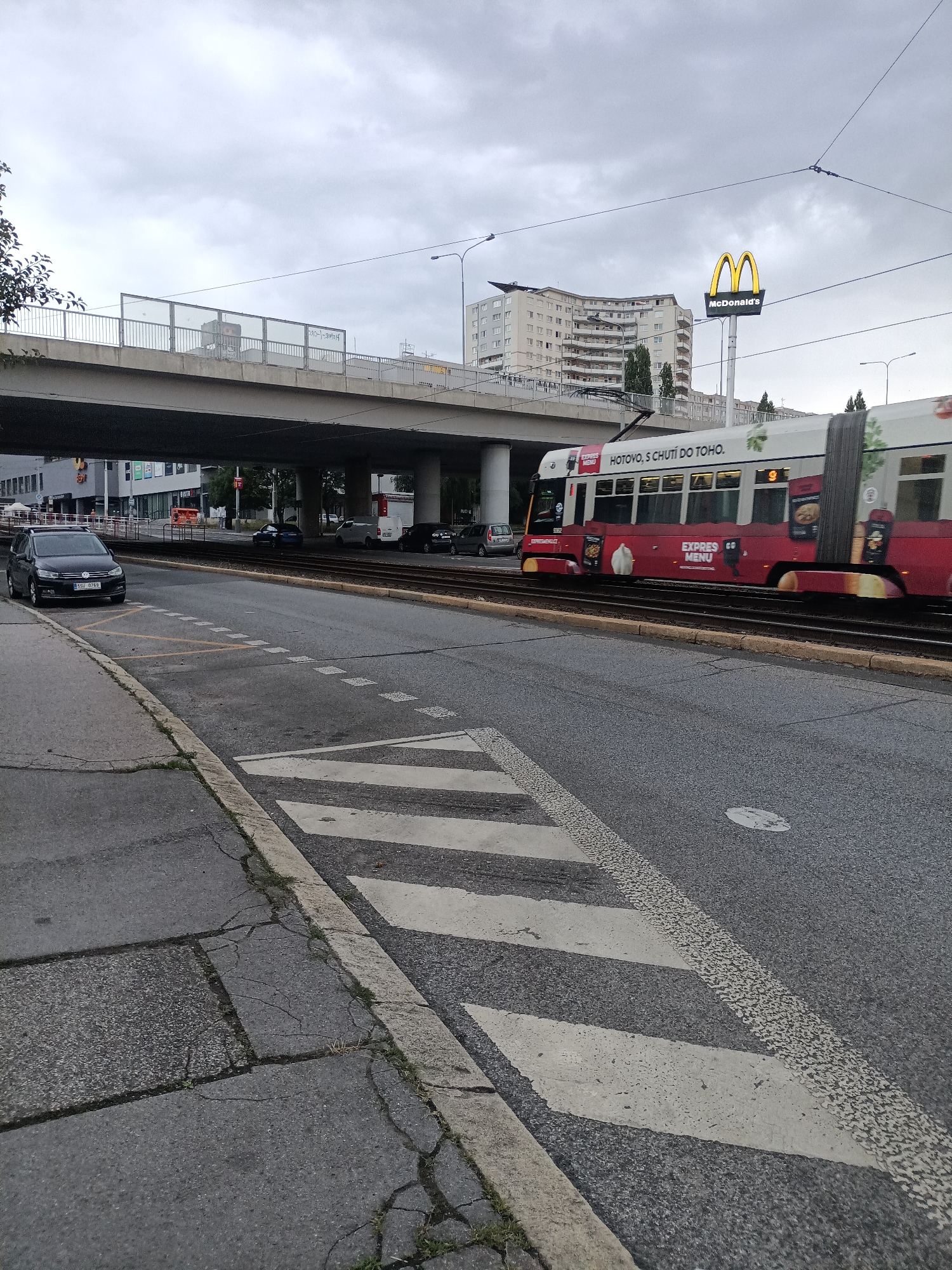 This is an exterior, eye-level, full shot of a street scene in Prague, Czechia. The photograph was taken on an overcast day. In the foreground, on the left side of the image, is a sidewalk with cracked concrete and a concrete curb. Next to the sidewalk is a street with tram tracks running through it. On the right side of the street is a crosswalk with white diagonal stripes.
In the middle ground of the image, a red and white tram is moving from left to right. The tram has advertisements on its side, including images of food and text that reads "HOTOVO, S CHUTÍ DO TOHO." and "EXPRES MENU." Further back, a dark grey SUV is parked on the left side of the street. Beyond the tram, several cars are visible on the street, including a white van and a dark sedan.
In the background, there is a large overpass or bridge structure with a metal railing and a glass barrier on top. Below the overpass, there are buildings with windows and signs. A large, illuminated McDonald's sign with its distinctive golden arches is visible above the buildings. A tall, multi-story apartment building with a curved facade is also prominent in the background. The sky is cloudy and gray.