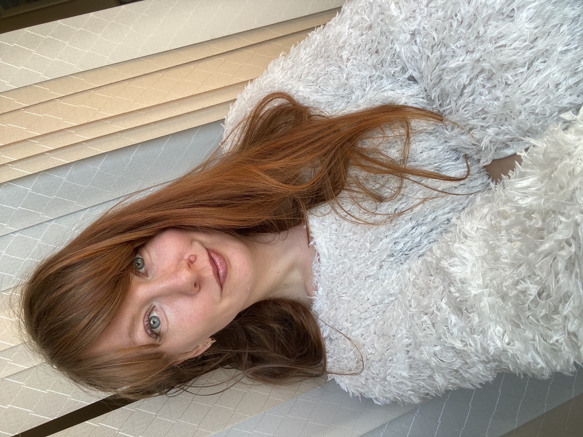 A woman with red hair and blue eyes lies on a surface of patterned beige and grey material. She is wearing a white, fluffy jacket. The setting is indoors, likely in a residential or commercial space in Krasnoyarsk, Russia. Based on the patterns visible in the background and foreground, the surface she is lying on appears to be composed of multiple layered blinds or textured panels, possibly samples of different wall or window coverings.