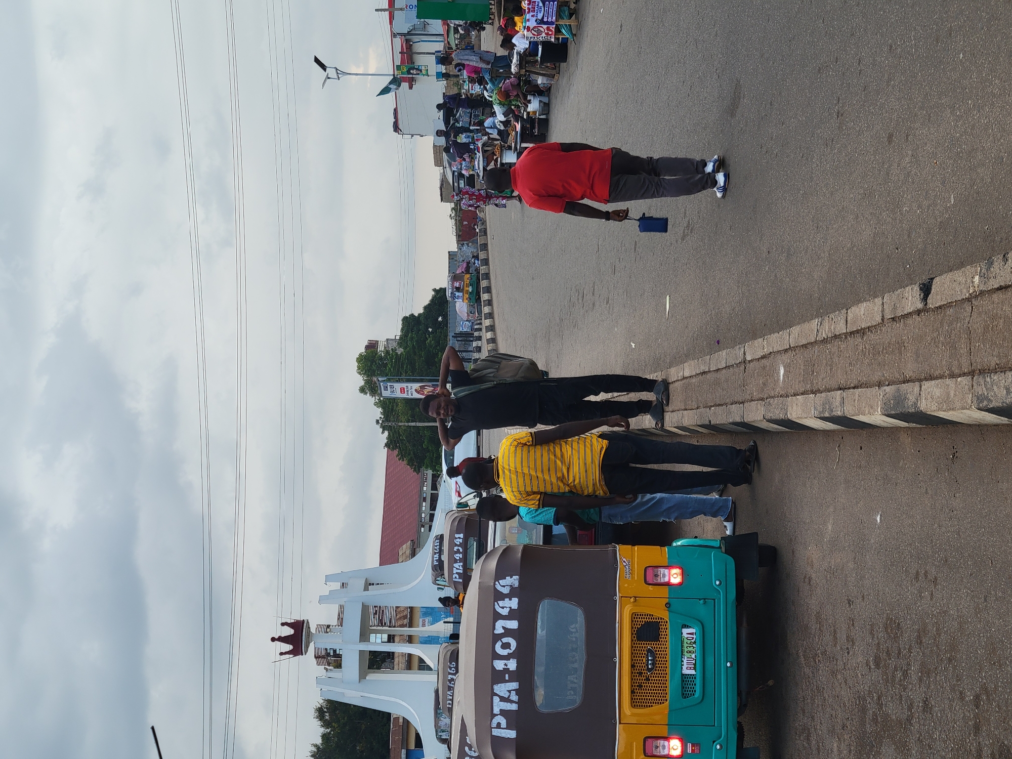 A street scene in Barakin Lerawi, Nigeria, captures a cloudy day with a monument featuring a crown at its apex in the background. Several "Keke Marwa" (auto rickshaws) are parked along the roadside, with one prominently displaying "PTA-10744" and a license plate "RUU-8360A". In the mid-ground, a group of men stand by the roadside. One man in a black t-shirt and dark pants has his hands behind his head and a bag slung over his shoulder. Another man in a red t-shirt and grey pants walks away from the camera, carrying a blue bag. Further in the background, various vendors are set up along the street, selling goods from tables. The road is paved, and a concrete curb with black and white stripes separates the road from a pedestrian area. The environment appears to be a busy urban setting.
