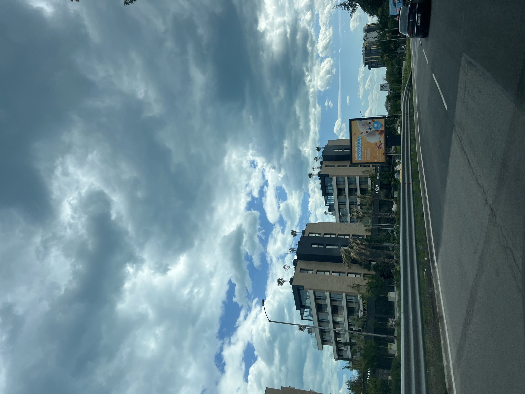 The image depicts a view from a vehicle on a road in Varna, Bulgaria. The road is flanked by modern buildings, including apartment complexes, visible against a sky of partly cloudy weather. A large advertising billboard is present along the roadside. A guardrail separates the road from the adjacent structures. One vehicle is visible driving on the right side of the road.