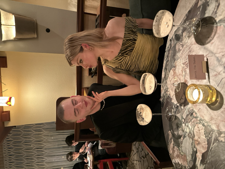 At a dimly lit restaurant, a blonde woman in a patterned top leans towards a man in a black suit and pearl necklace. Three champagne glasses and a candle holder are on the marble table. A small card reads "Sorry, I'm Taken." Behind them, other diners are visible. The background includes patterned wallpaper, wooden structures, and warm lighting. The overall atmosphere appears upscale and intimate.