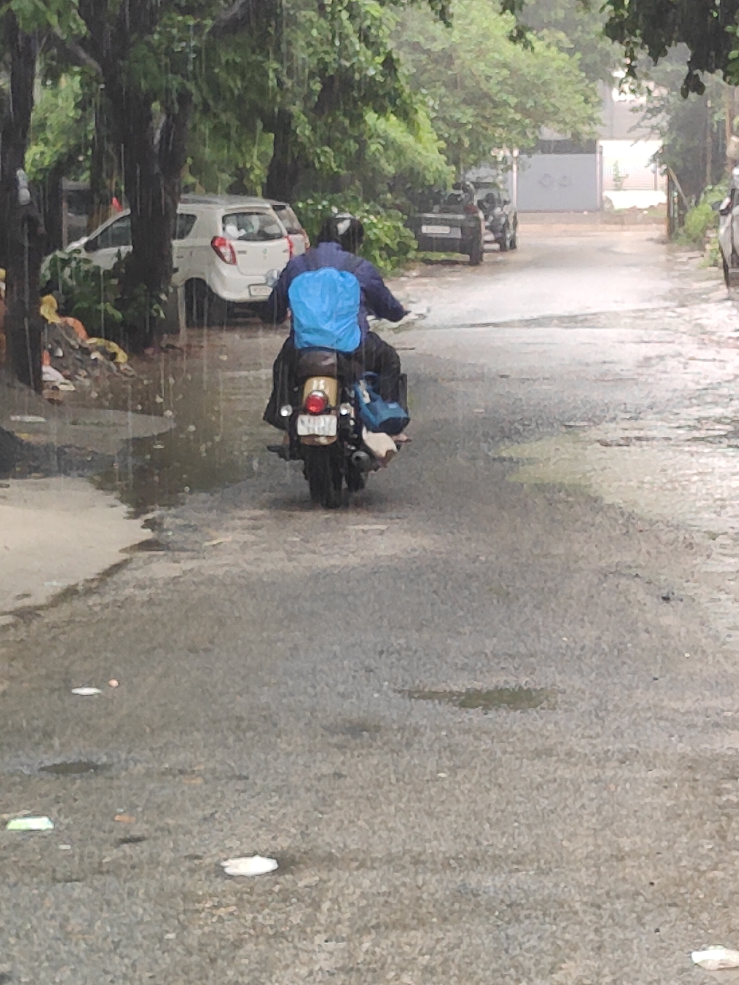 The image shows a person riding a motorcycle down a wet, asphalt road in Gurgaon, India, during a rainstorm. The rider is wearing a blue poncho and a helmet, and is carrying luggage on the back of the motorcycle. The road is lined with trees and parked cars. The scene appears to be a residential or commercial area, with buildings visible in the background. The overall atmosphere is one of overcast weather and a moderate downpour.