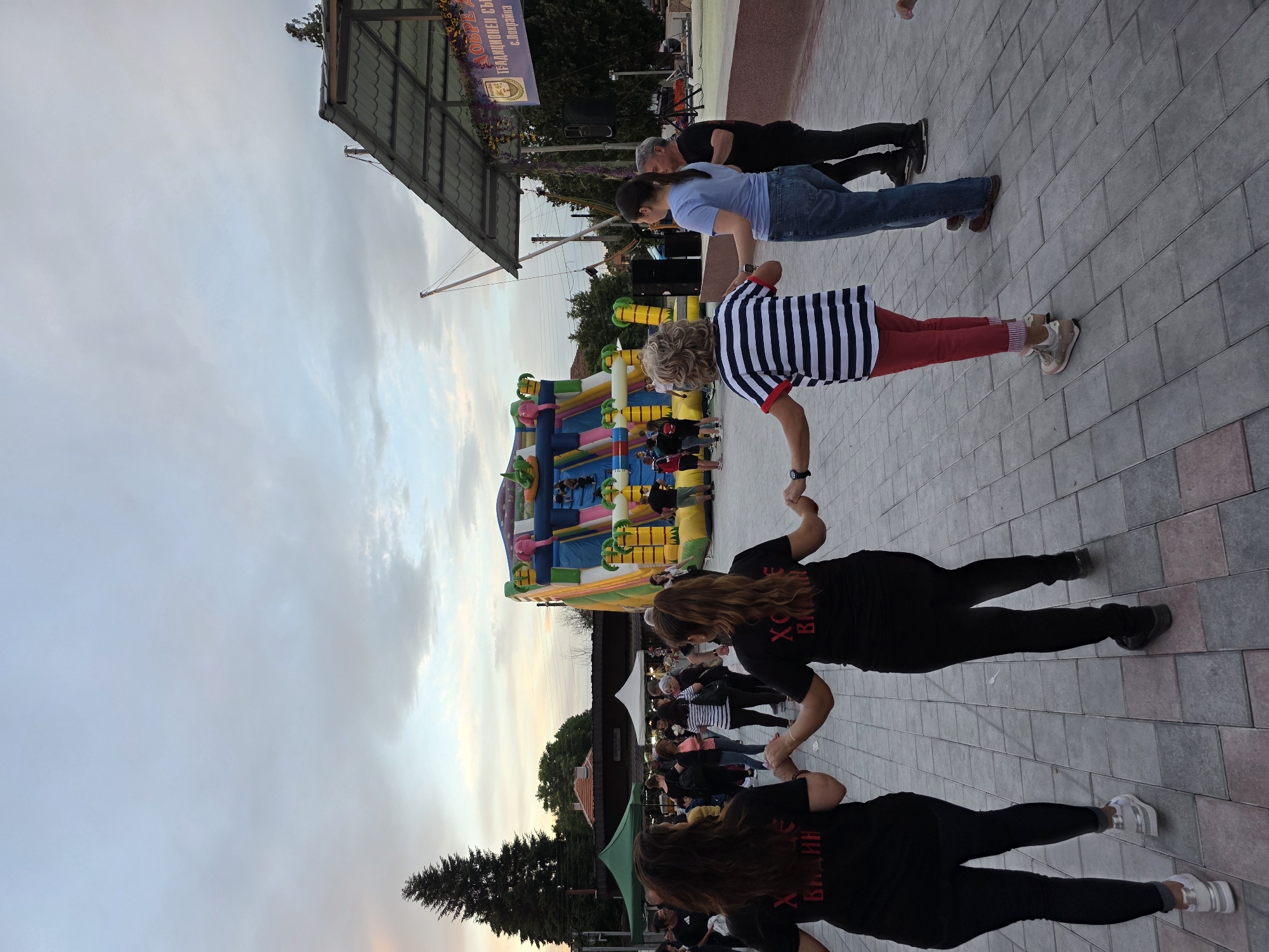 The image shows a group of people performing a traditional Bulgarian folk dance in Pokrayna, Bulgaria. The dancers, in a line, are holding hands. Some wear black shirts with Cyrillic text on their backs; another person wears a striped shirt and red pants, while a third wears a blue shirt and jeans. A bounce house is visible in the background, along with a crowd of onlookers. The setting appears to be an outdoor event, taking place on a paved square with buildings and trees in the background under a cloudy sky. A stage structure with signage in Cyrillic is also present.