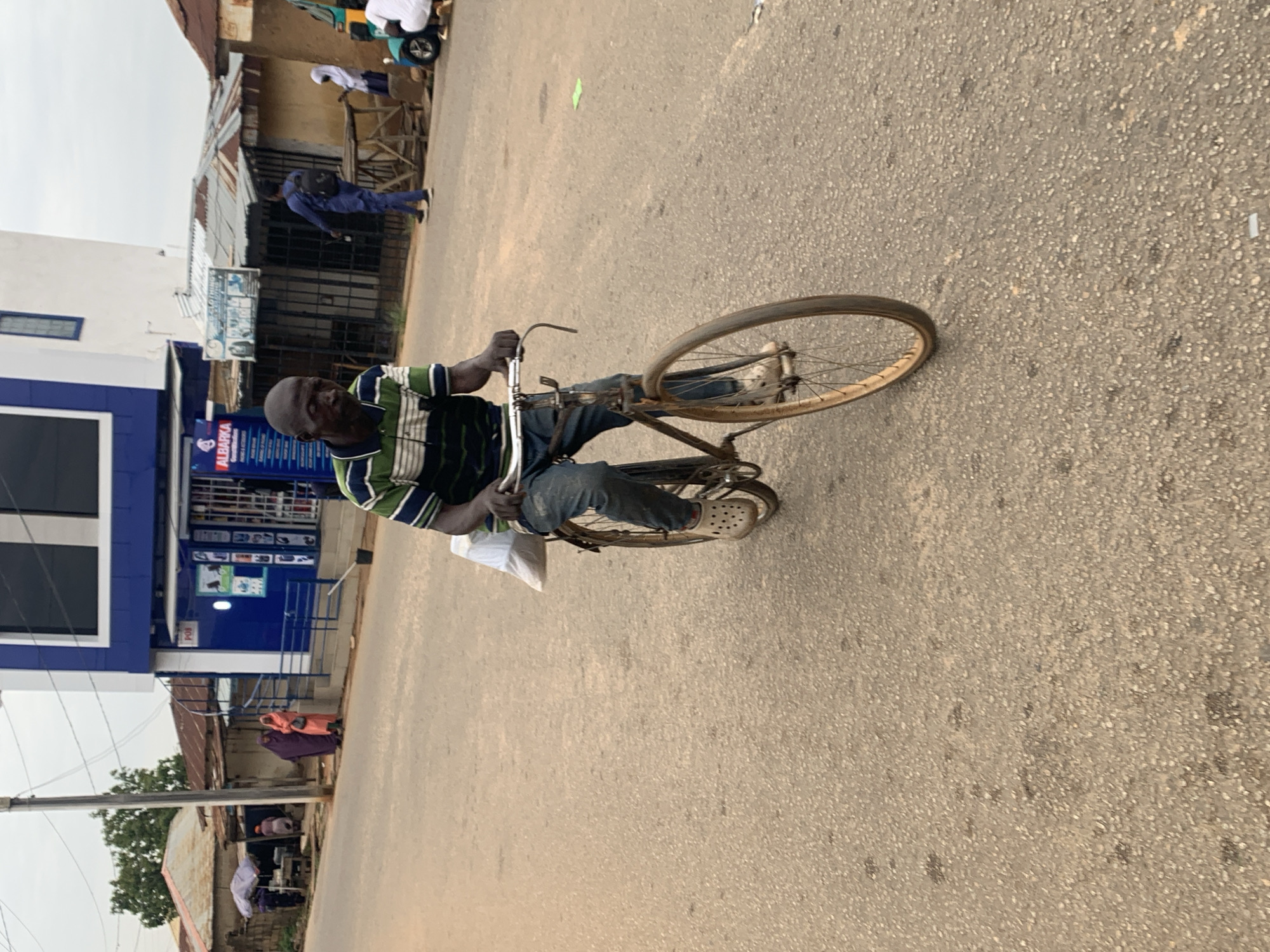 A man is riding a bicycle on a paved street in Jos, Nigeria. He is wearing a green and blue striped polo shirt, blue jeans, and tan Croc-style shoes. A white plastic bag is strapped to the back of his bicycle. In the background, there is a blue and white building with signs in English and what appears to be Arabic script. Several other people are visible walking and standing on the sidewalk, as well as a blue tricycle and a white vehicle. The sky is overcast.