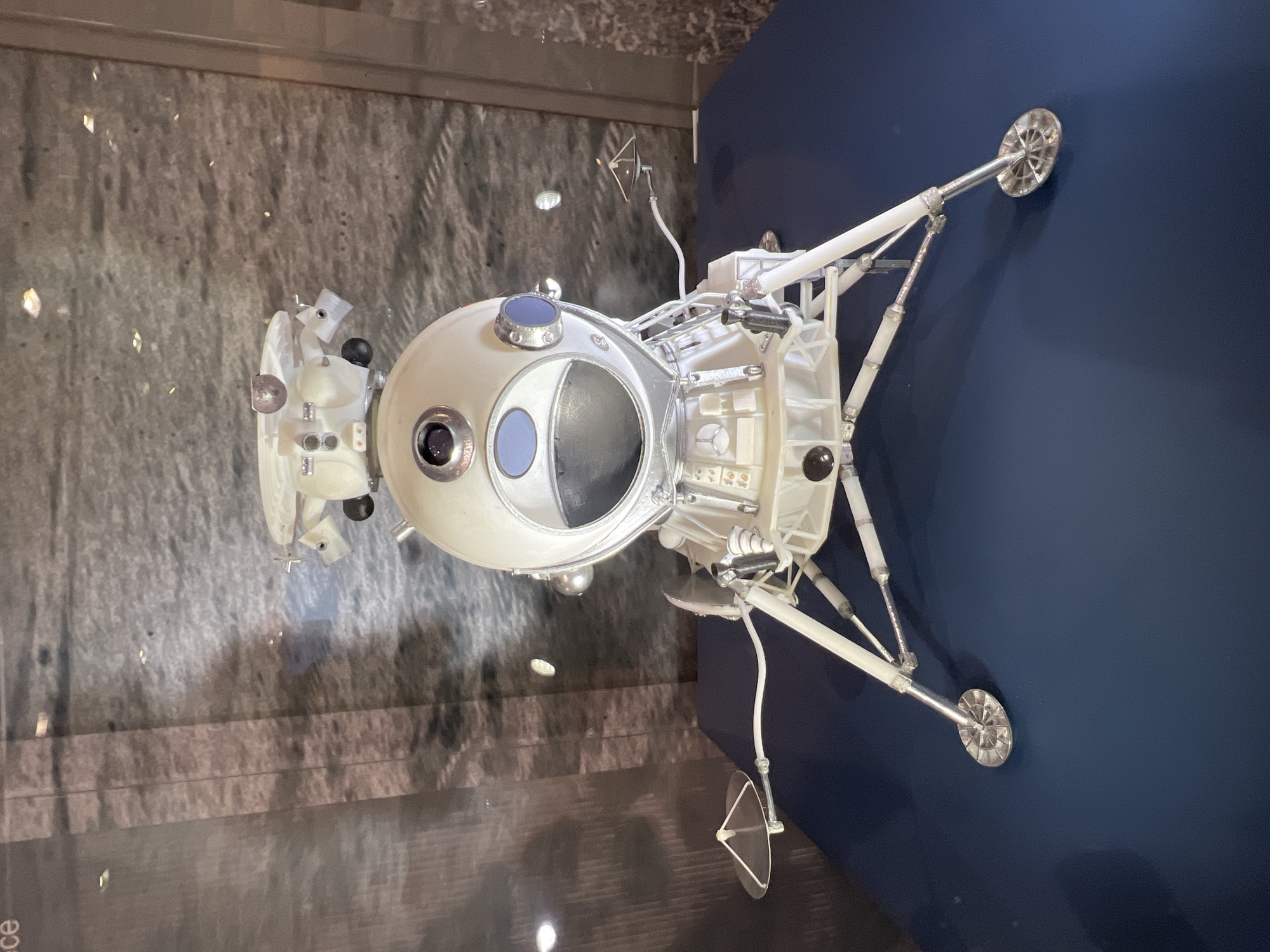 The image shows a model of a Luna 16 lunar lander, likely on display in a museum. The lander has a spherical upper section with a large circular opening, possibly for sample collection or instrument placement. It has three landing legs with circular pads at the bottom. The background consists of a textured wall on one side and a smooth, dark blue surface on the other. This could be the State Museum of the History of Cosmonautics, named after K. E. Tsiolkovsky, in Saint Petersburg, Russia.