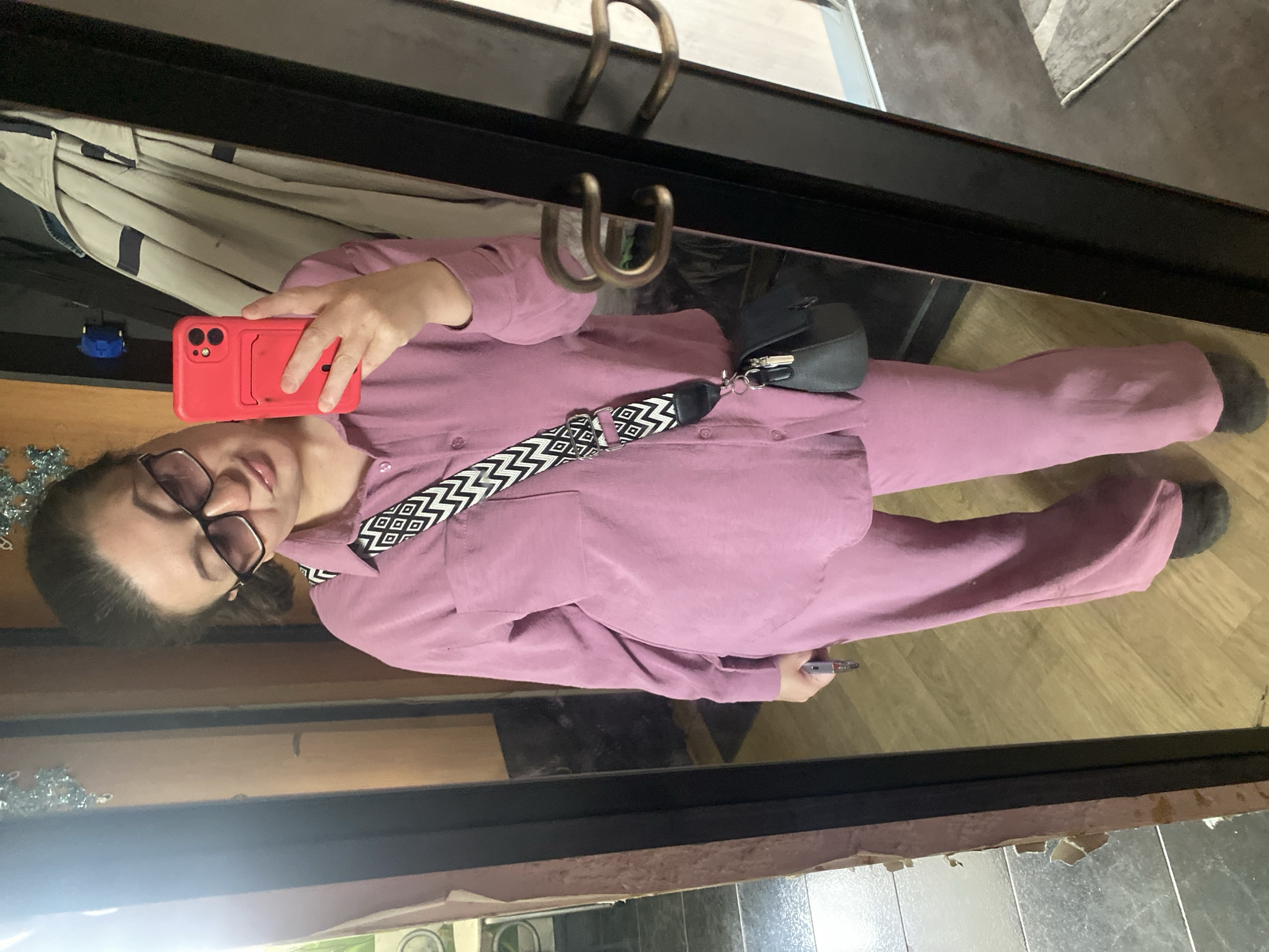 A woman stands in front of a mirror, taking a selfie with a red phone. She is wearing a matching lilac pajama set, a bag with a black and white strap, and glasses. Her hair is pulled back. The reflection shows parts of the room, including flooring, a coat hanging on a rack, and the trim of the mirror. It is impossible to determine whether this image was taken in Chaikovsky, Russia based on visual information.