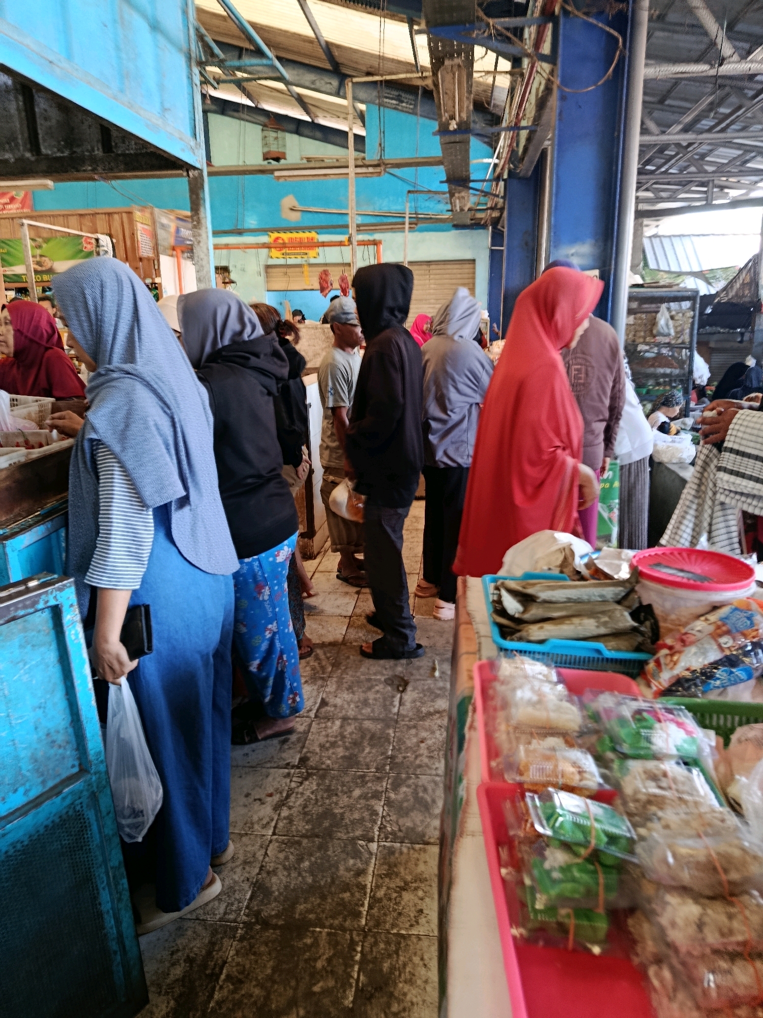 The image depicts a busy indoor market scene, likely in Bululawang, Indonesia. In the foreground, a person wearing a bright red hijab is partially visible, facing a stall. Next to them, a person in a grey hijab is also present. Behind them, several individuals are browsing or standing, some in dark hooded tops, others in casual attire. The market stalls are laden with goods, including packaged food items in pink and green plastic trays and what appear to be dried fish or similar items in a blue plastic basket. The environment is functional, with exposed metal beams and blue painted structures overhead, characteristic of a traditional market setting. The floor is tiled and shows signs of wear. Several bags, including a white plastic bag being held by a person in a blue outfit and grey hijab, are visible, suggesting active commerce. The overall impression is one of a bustling local market with various food products for sale.