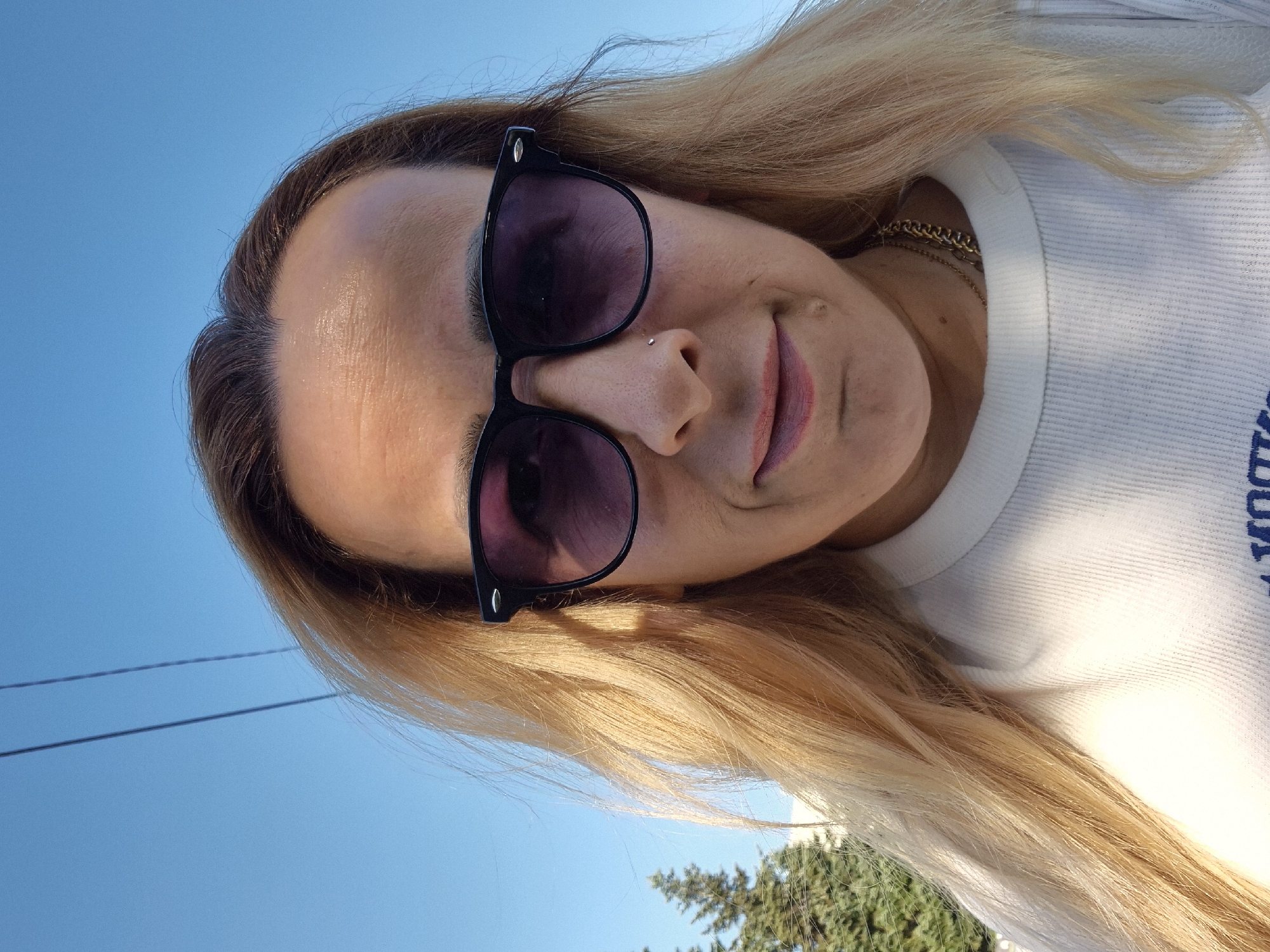 A low-angle, close-up selfie shows a woman from the chest up, wearing sunglasses and a white ribbed t-shirt. She has blonde hair that falls around her face, and is wearing a gold chain necklace. The background is a bright blue sky with two dark lines, possibly power lines, running across it. To the left of the woman, a portion of a tall green tree is visible. The image appears to have been taken outdoors on a sunny day.