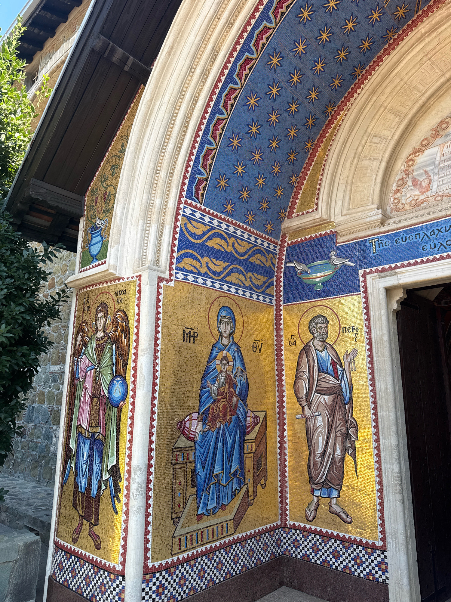 The image shows an exterior view of the Holy Cross Monastery in Tsakistra, Cyprus. The scene focuses on a section of the monastery's facade adorned with colorful mosaics. On the left, a mosaic depicts an angel with wings, holding a blue orb. To its right, a mosaic of the Virgin Mary holding the infant Jesus is shown. Further to the right, a mosaic features a man, likely Saint Peter, holding keys and a scroll. Above these figures, decorative mosaics with geometric patterns, stars, and stylized waves fill the archway. A small mosaic of a blue vase with a dove is visible above the angel. The architecture includes stone walls and wooden eaves. The overall impression is of religious art integrated into the building's exterior.