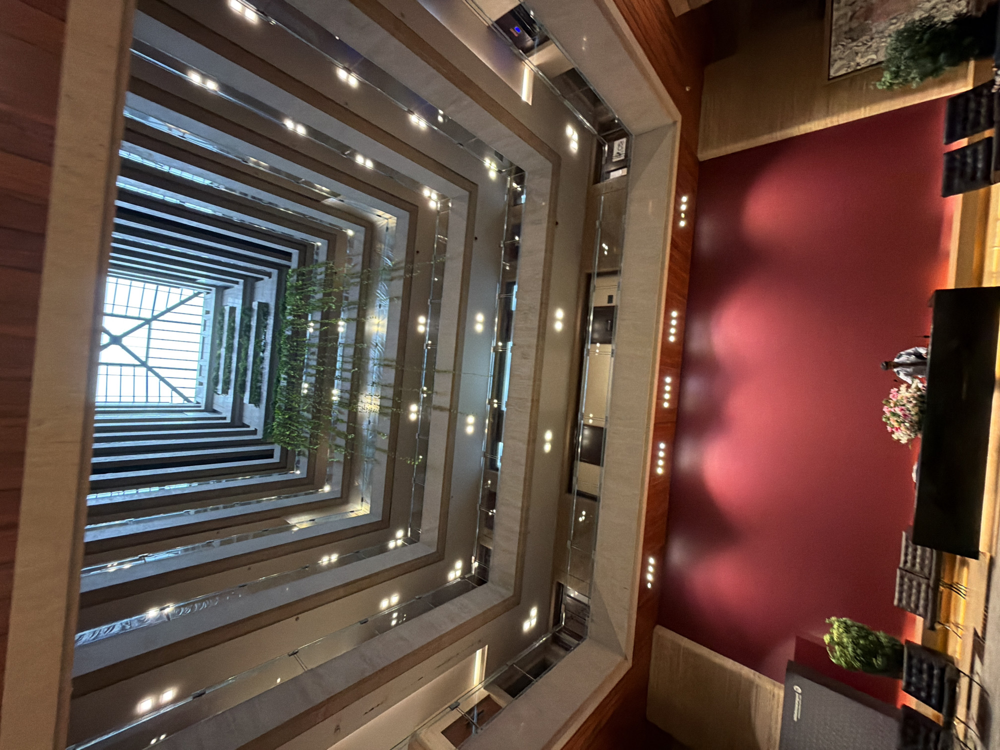 This is a wide-angle, low-angle interior shot looking up at a multi-story atrium. The atrium features stacked, cantilevered floors with glass railings, creating a tiered effect. Suspended plants hang from some of the upper levels. A skylight is visible at the very top of the atrium. The lower portion of the image shows a reception area with a red wall, several dark chairs, and a desk. A person in a light-colored shirt and dark pants stands behind the desk, and a floral arrangement is on the desk. A large, dark, angular structure is to the left of the desk. The lighting is artificial, with numerous small lights visible along the edges of the floors. The architectural style appears modern, with a focus on clean lines and geometric shapes. The image does not provide any direct clues about the specific city or country, including Mumbai, India.