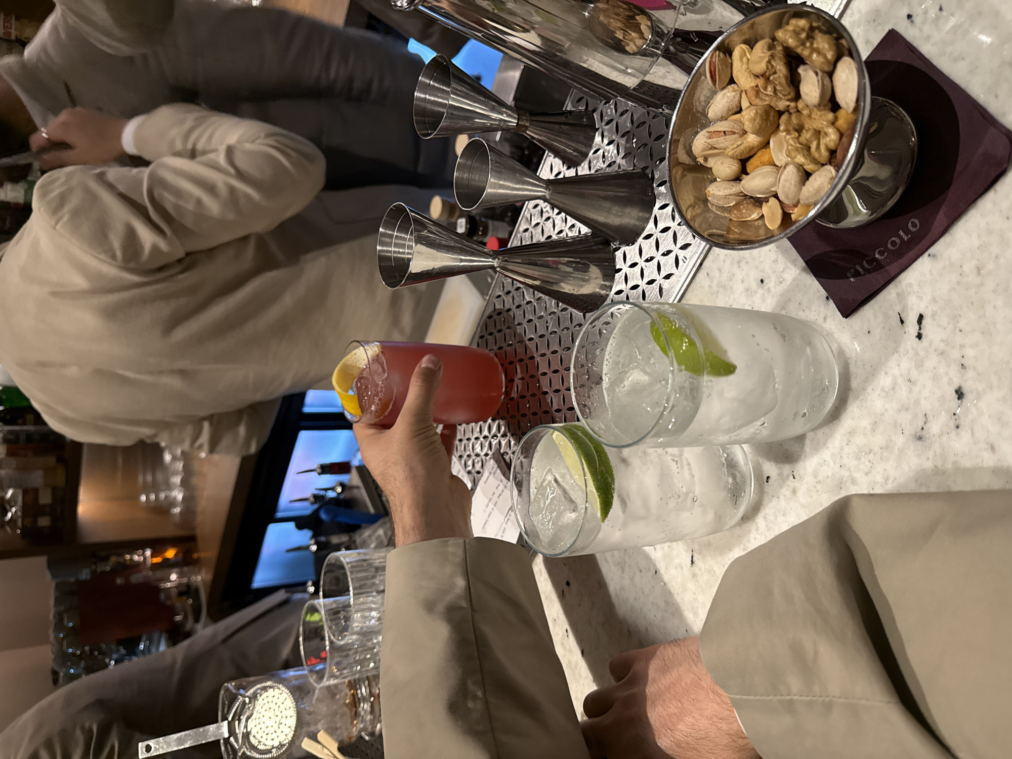 The image shows a bar scene with two people visible. One person in a light-colored suit is holding a cocktail with a lemon twist. Two other cocktails with lime wedges are on the bar. A silver container of mixed nuts rests on a dark purple napkin with "PICCOLO" printed on it. Three stainless steel jiggers are also on the bar. The bar top has a textured white surface. The location is in Yerevan, Armenia.