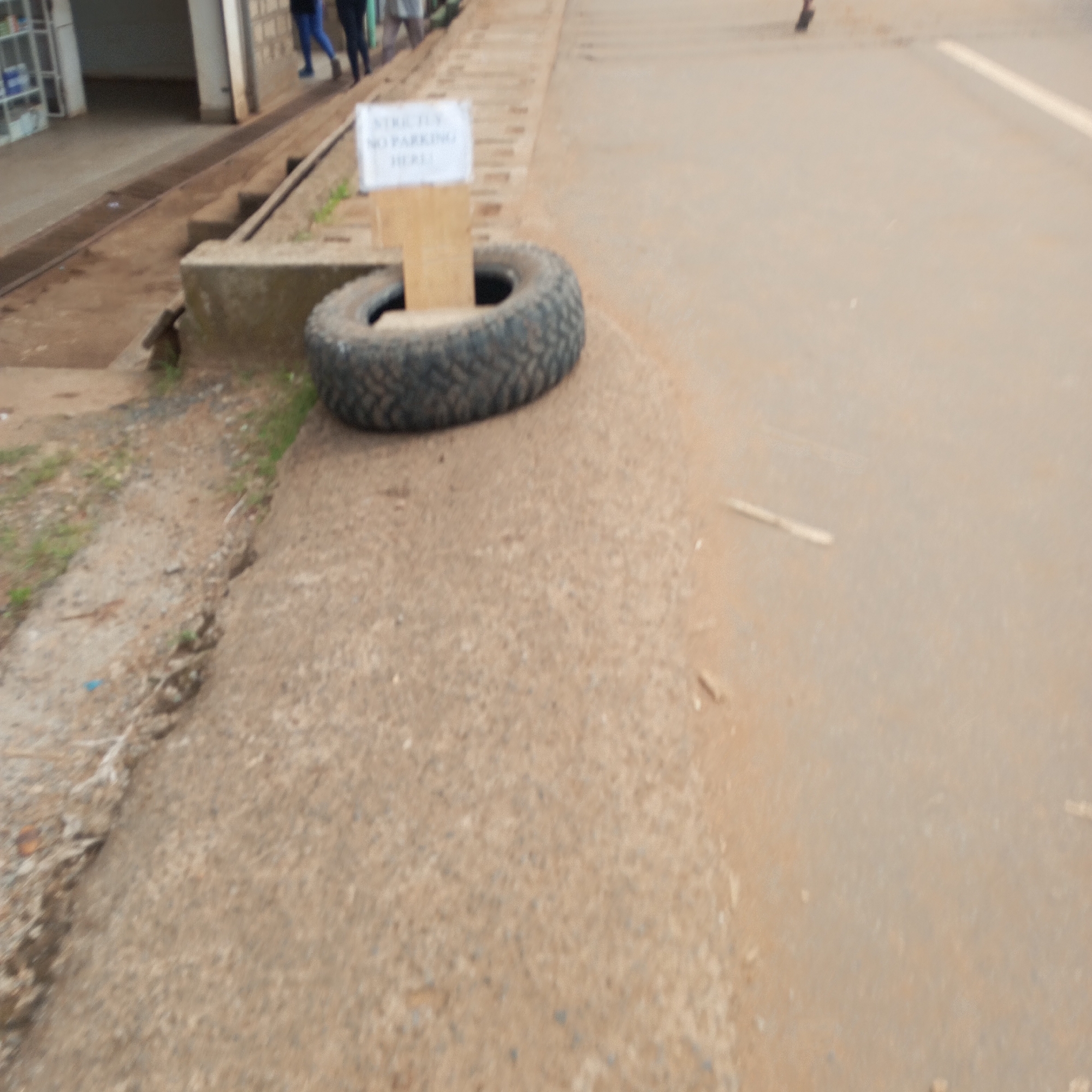 The scene shows a dirt and paved roadway in Sheywe ward, Kenya. A "STRICTLY NO PARKING HERE" sign is propped up by a tire on the edge of the road near a sidewalk. Several people are walking in the background.