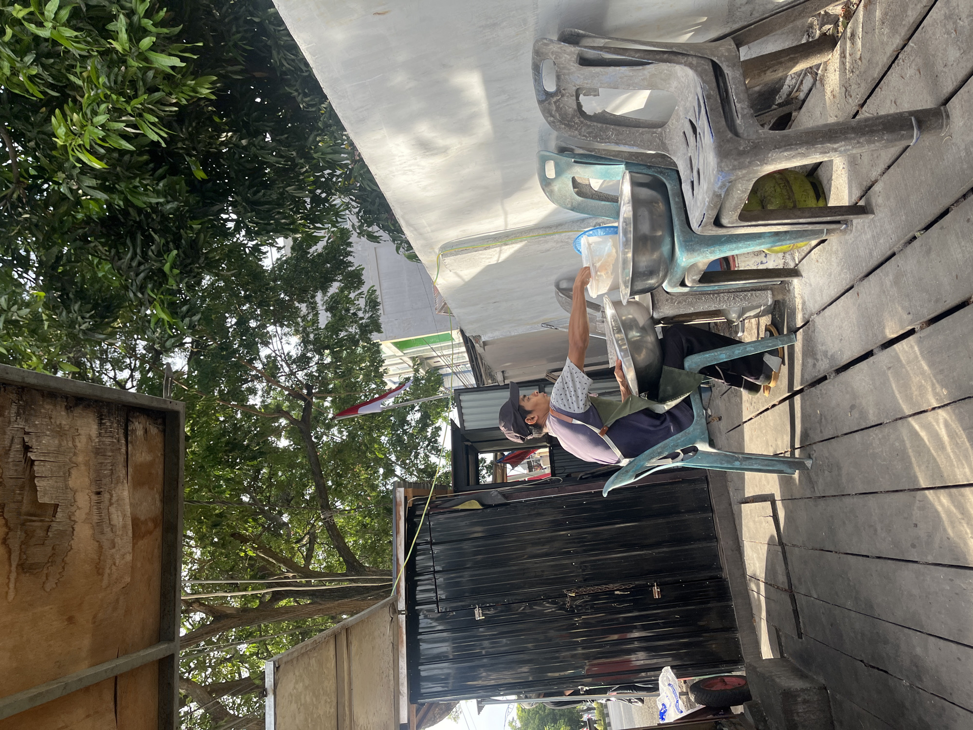 A man wearing a dark baseball cap and an apron is preparing food at a stall in Banda Aceh, Indonesia. He is seated on a blue plastic chair and is working with two large metal bowls and a white plastic container. Behind him, a black corrugated metal structure serves as the back of the stall. To the right of the stall, several stacked plastic chairs are arranged against a white concrete wall. The ground is made of weathered wooden planks. Lush green trees and a portion of a building with a red and white flag are visible in the background. The scene appears to be outdoors, with sunlight casting shadows on the ground, suggesting it is daytime.